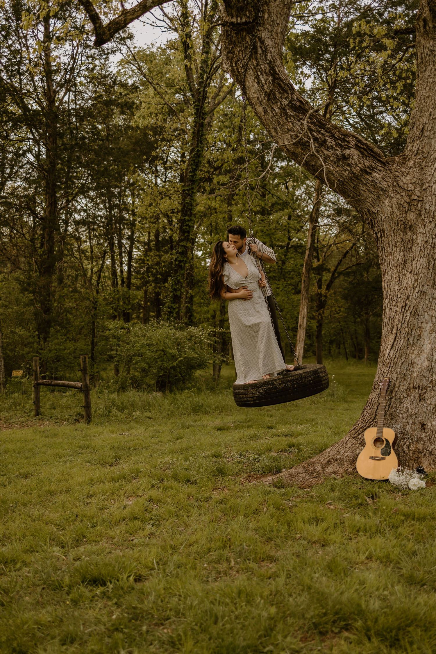 A couple embraces on a tire swing in a rustic outdoor setting with a guitar nearby.