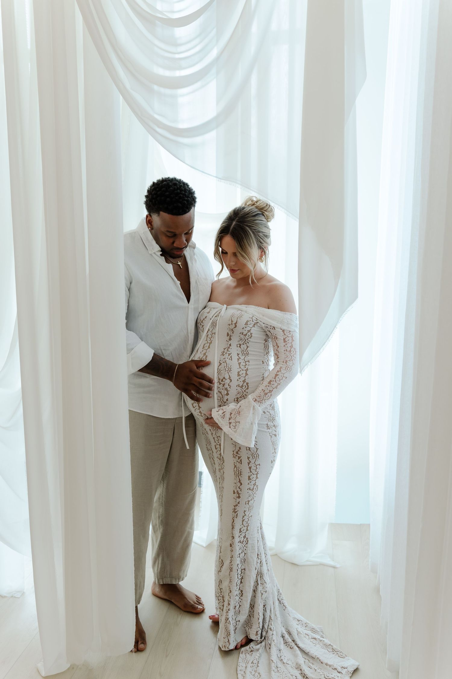 A bride in a white lace dress and groom in light colored suit embrace in front of flowing white curtains.