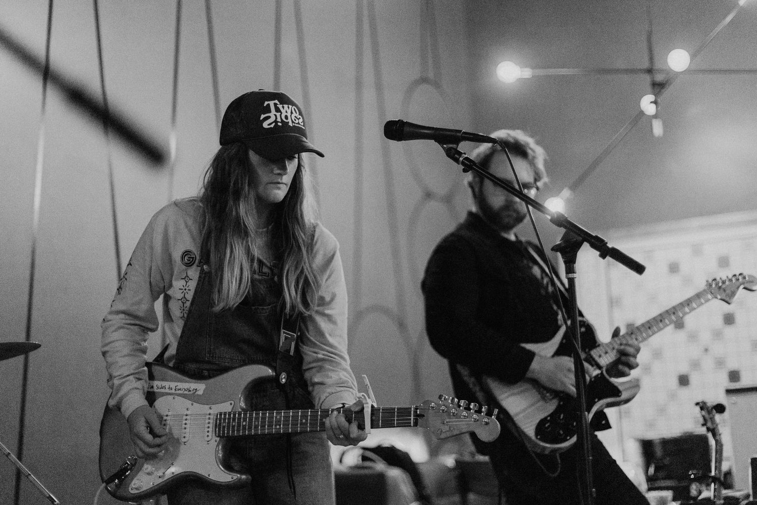 Musicians perform with guitars in a monochrome setting with stage lighting.