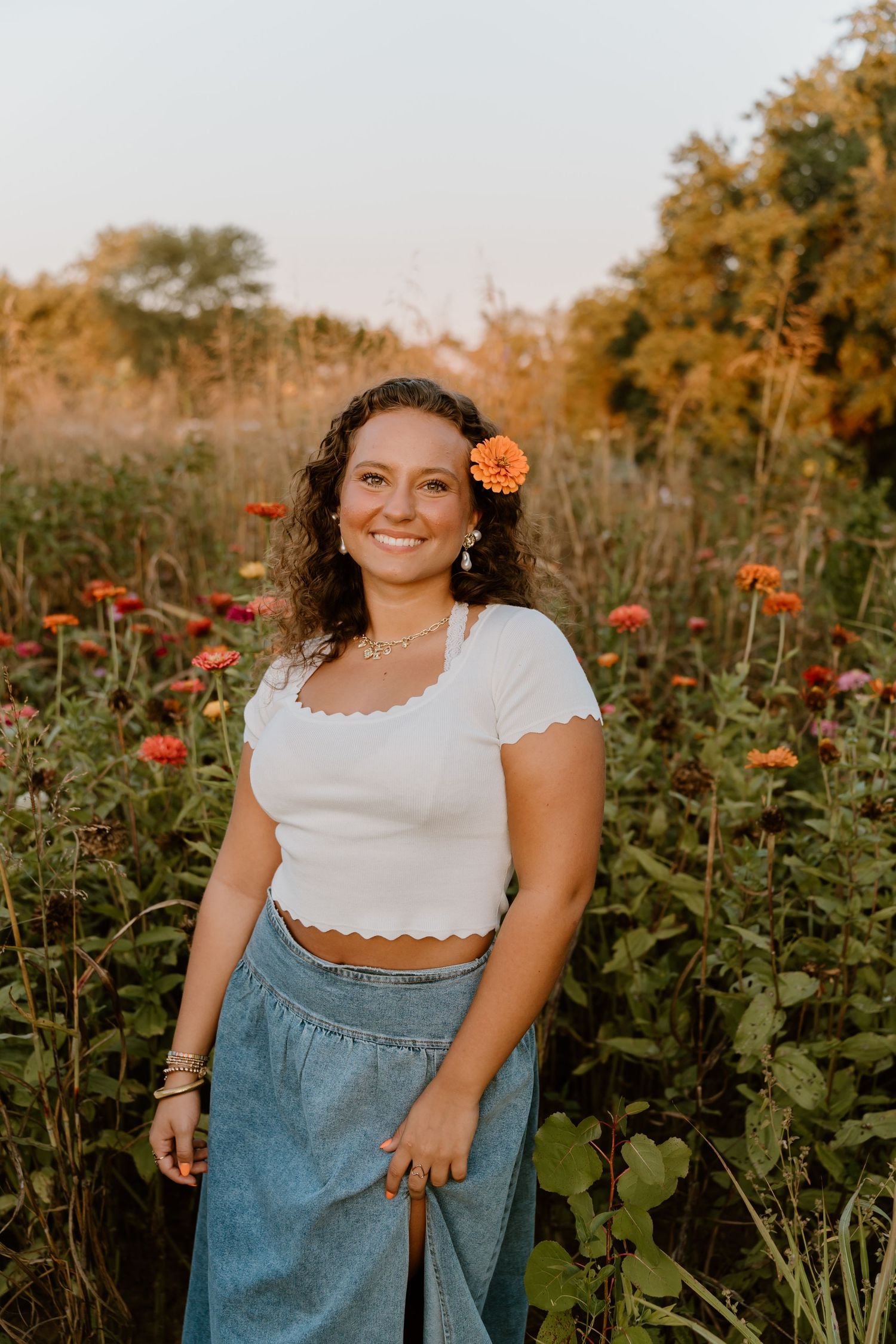 Senior girl with orange flower in hair poses in white top and denim among blooming zinnias.
