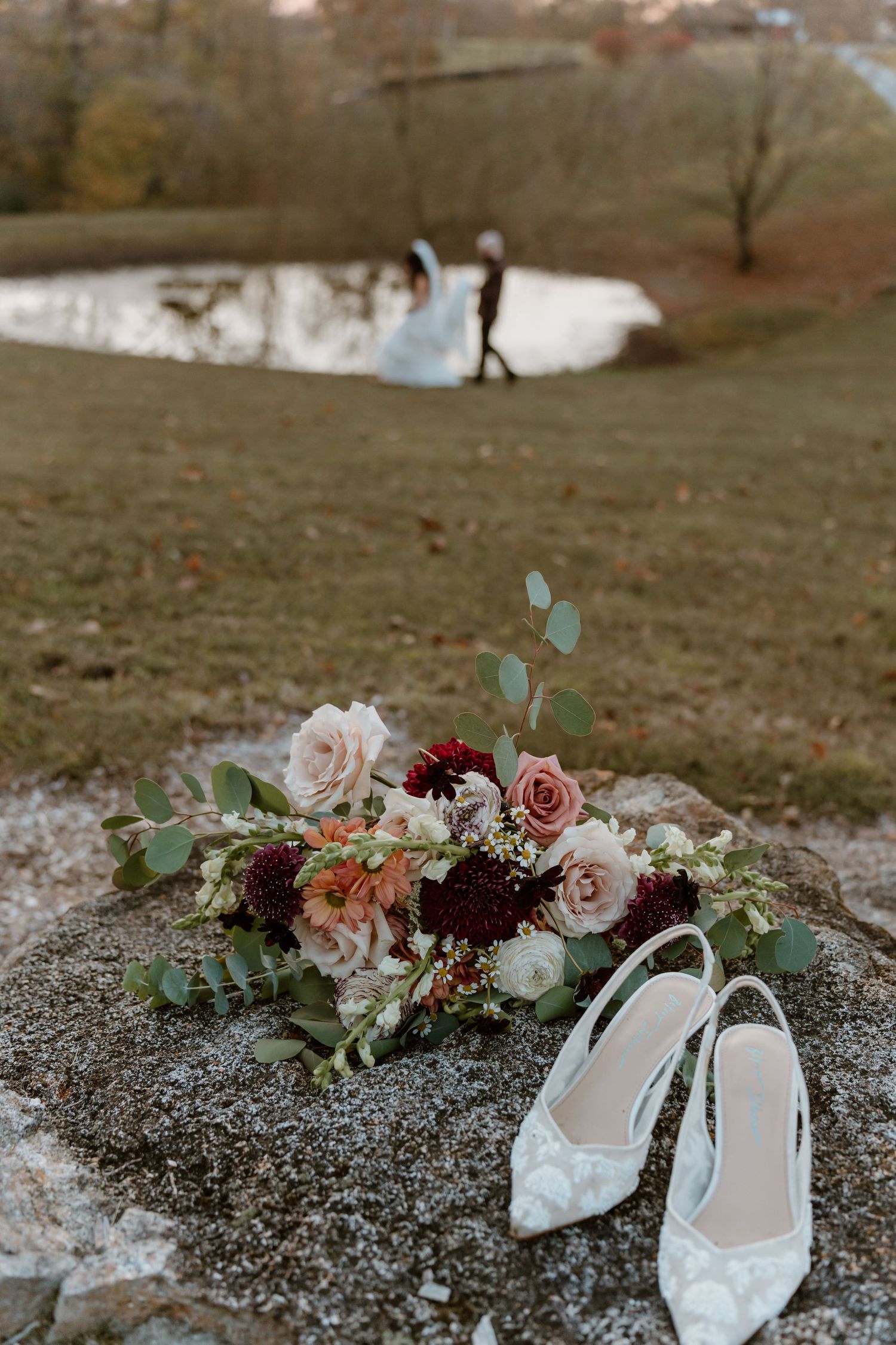 White lace bridal shoes and pink floral bouquet with burgundy roses arranged on rocky ground outdoors.