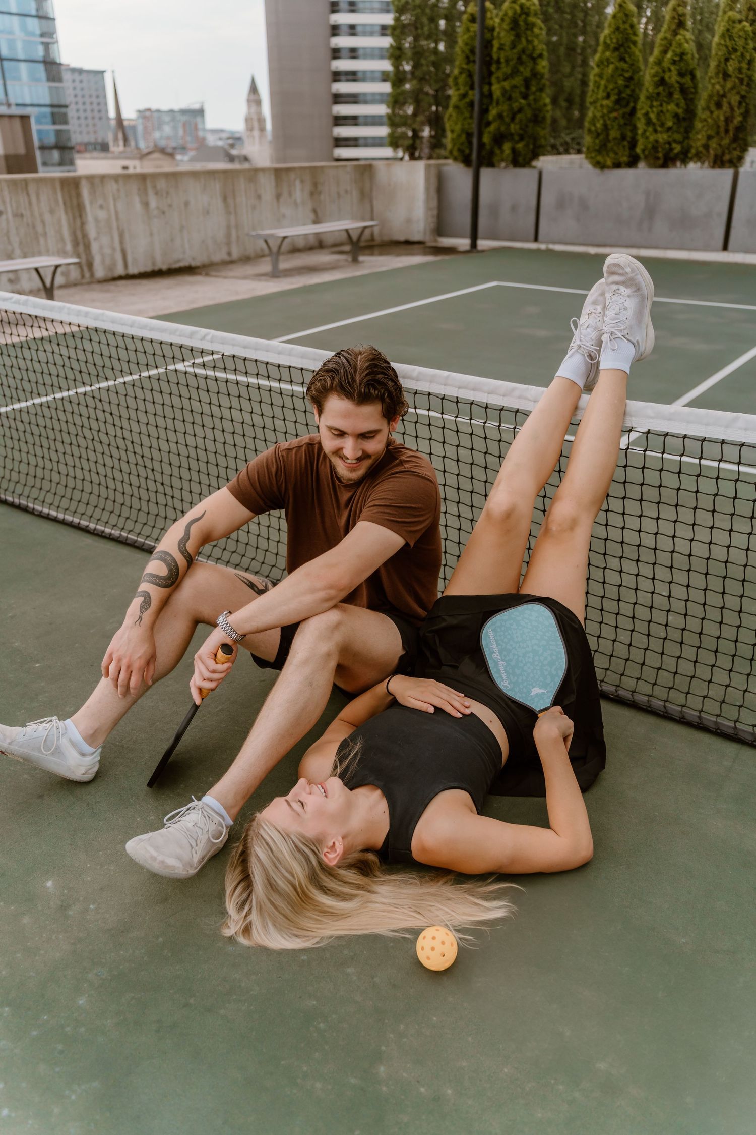 A couple laughs while lying on a tennis court with an orange ball.
