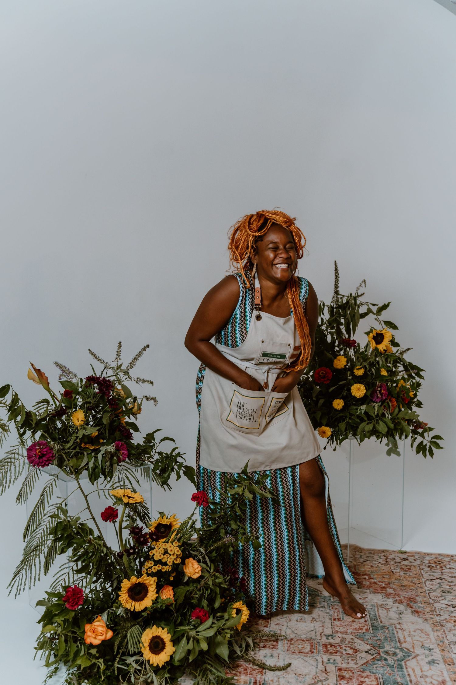 Person in striped dress stands joyfully among vibrant floral arrangements with sunflowers.