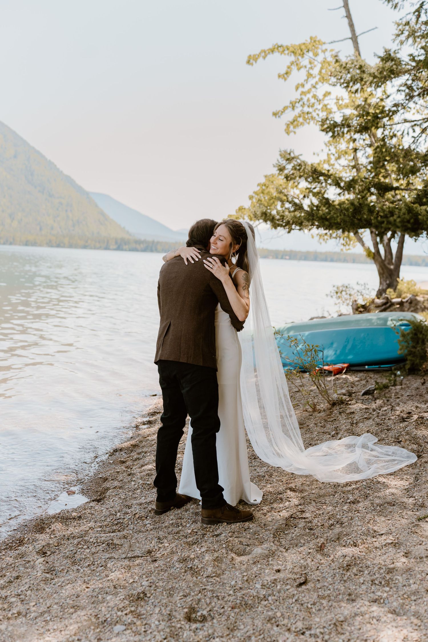A couple shares an intimate moment by a lake with mountains in the background and a blue canoe on the shore.