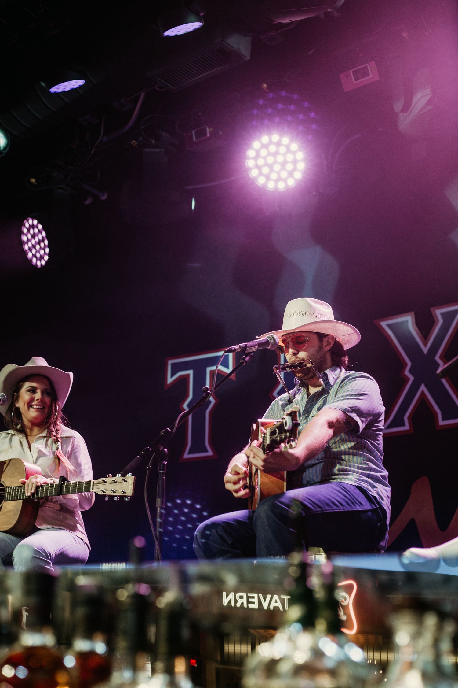 Musicians with acoustic guitars perform under purple stage lights at a concert venue.