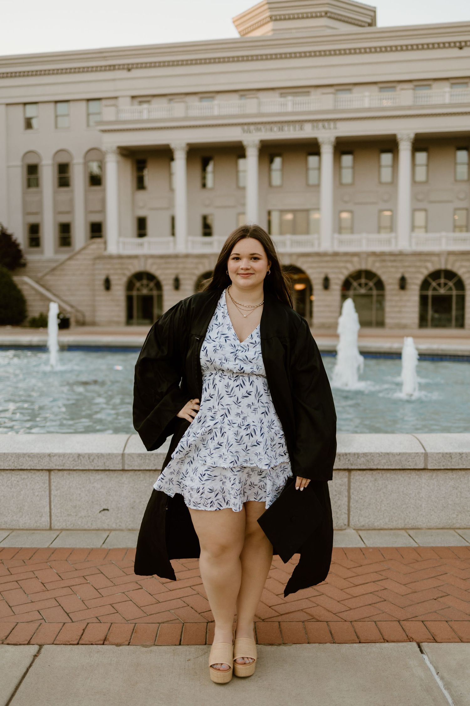Graduate poses confidently in floral dress and academic gown by ornate building and fountains.