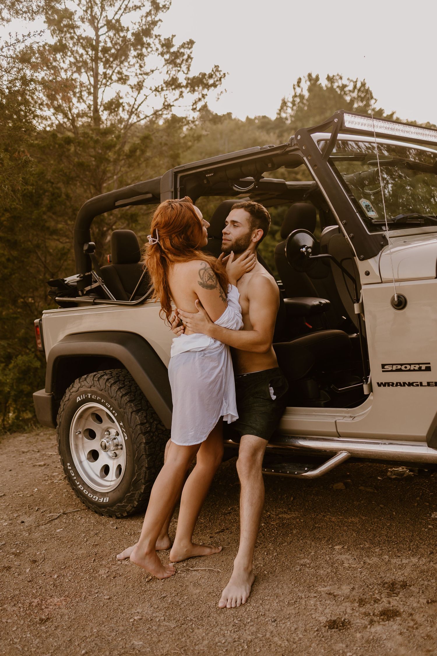 A couple shares tender moments by a Jeep Wrangler parked on dirt ground at sunset.