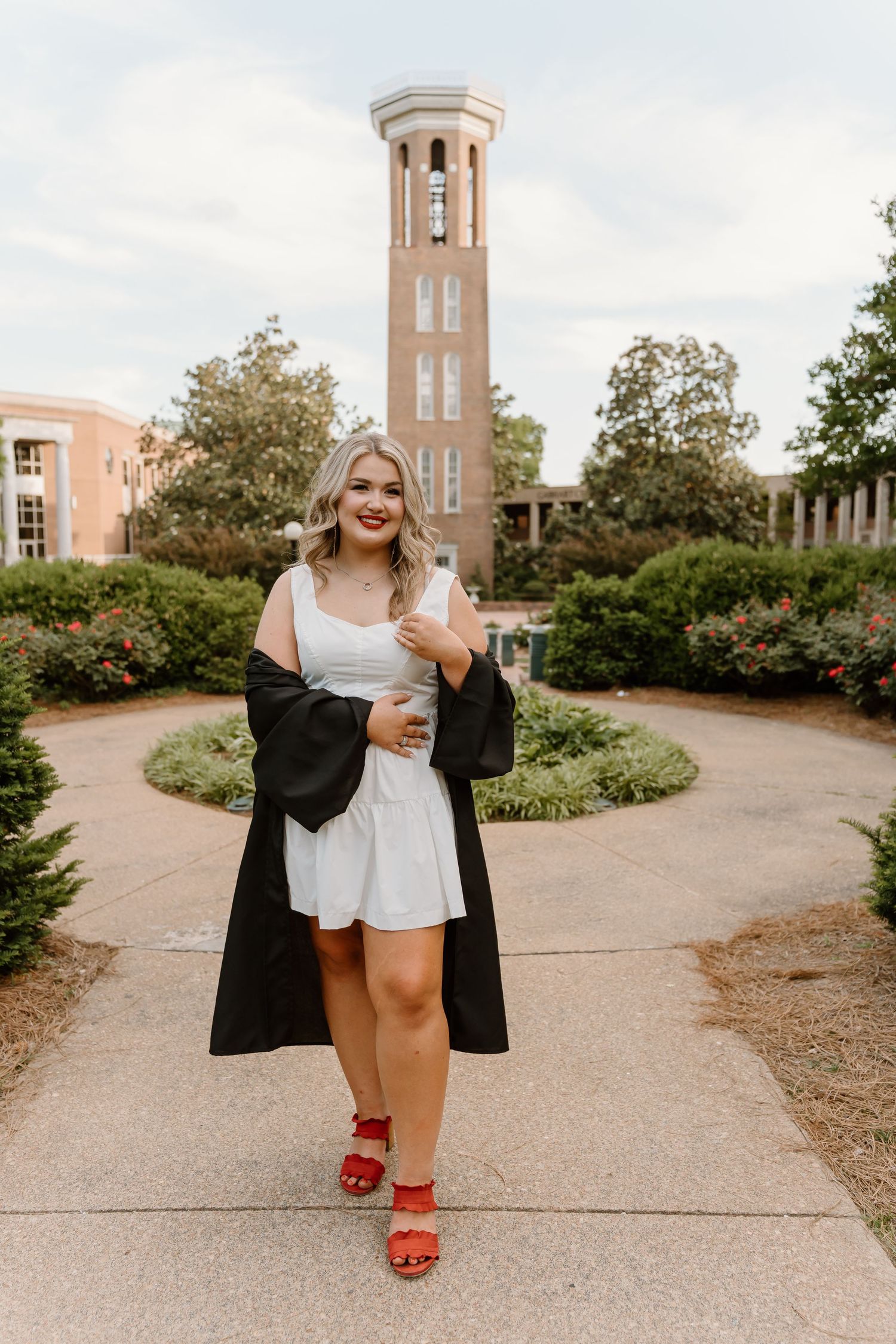Young woman in white dress and black graduation robe poses on campus pathway with brick bell tower backdrop.