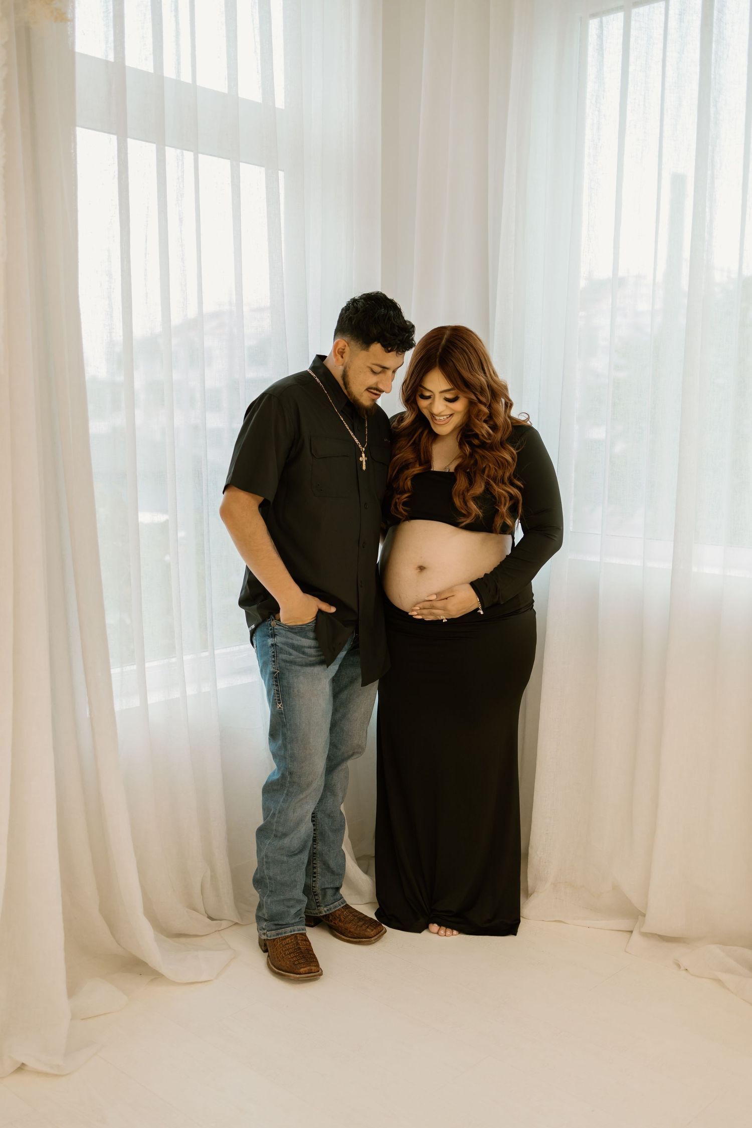 An expecting couple shares an intimate moment by sheer white curtains in a bright room.