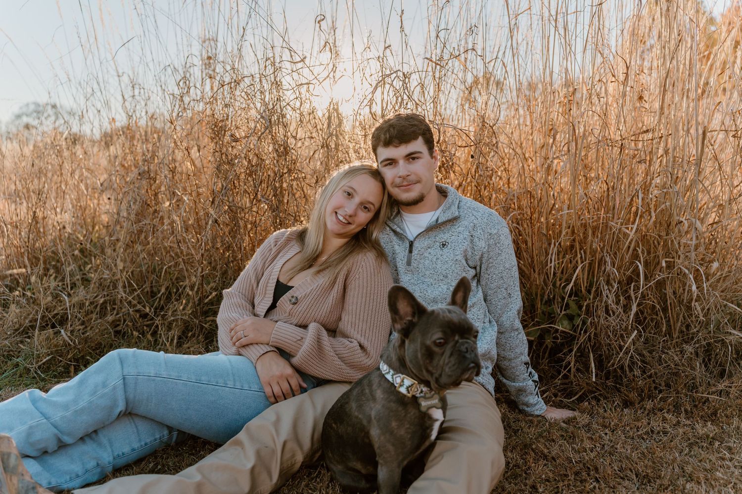 Couple in casual fall outfits sitting together in golden field during sunset photoshoot.