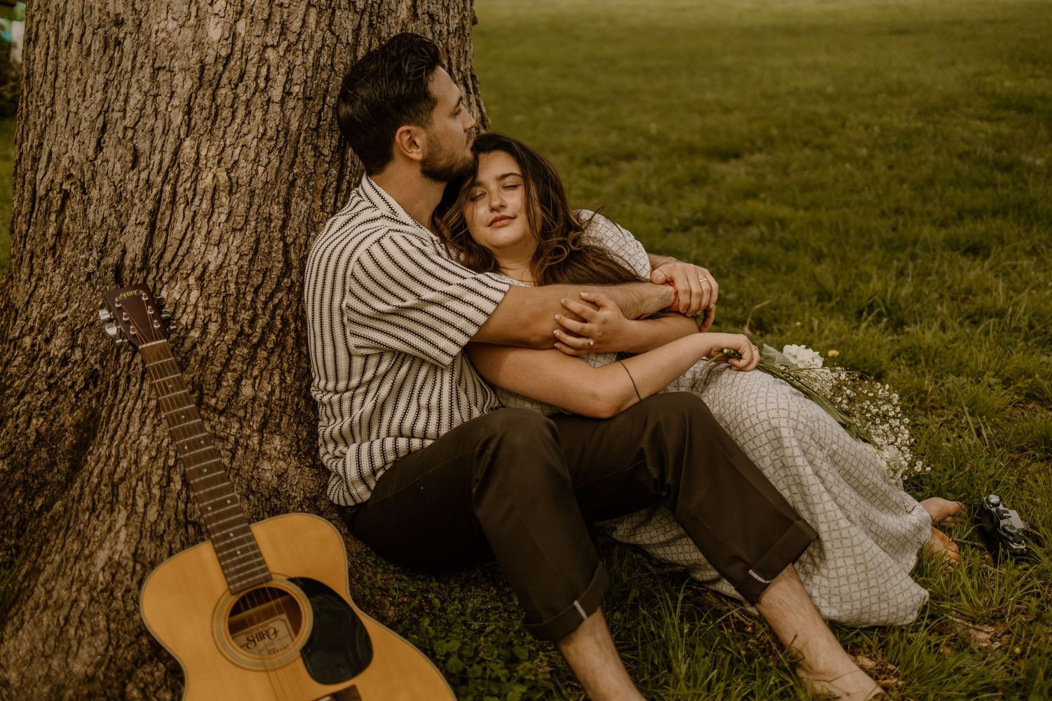A couple relaxes under a tree with a guitar during a romantic outdoor photoshoot.