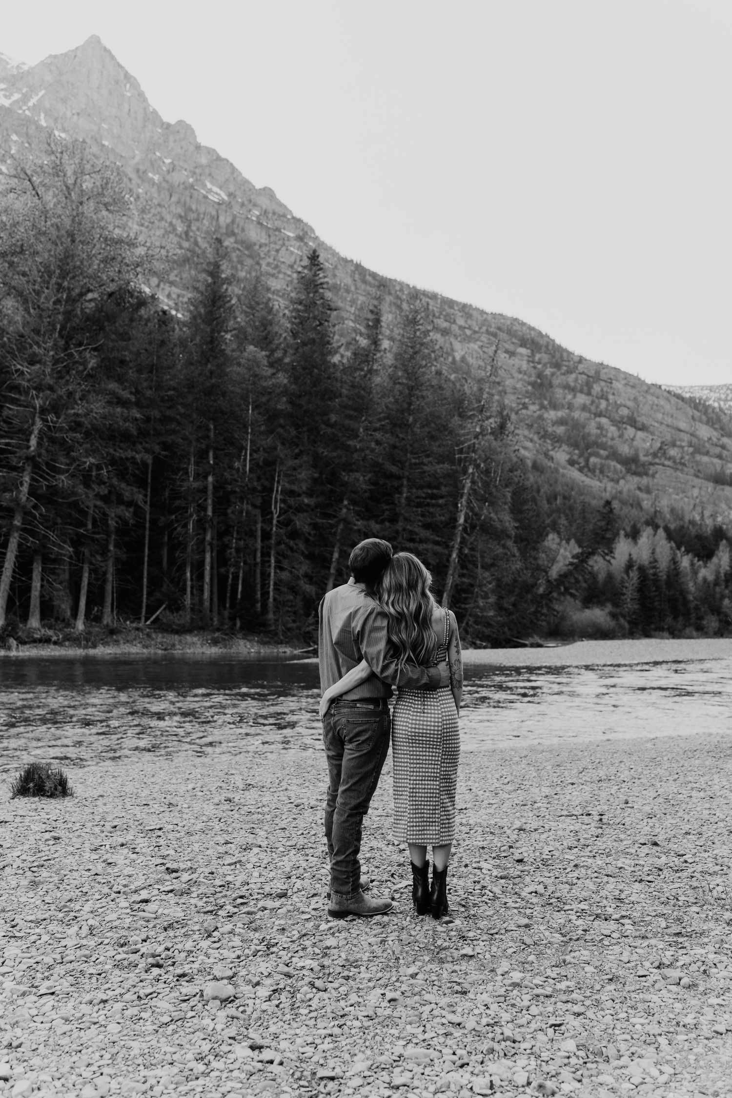 Two people walk along a rocky riverbed with mountain peaks and evergreen trees in the background.