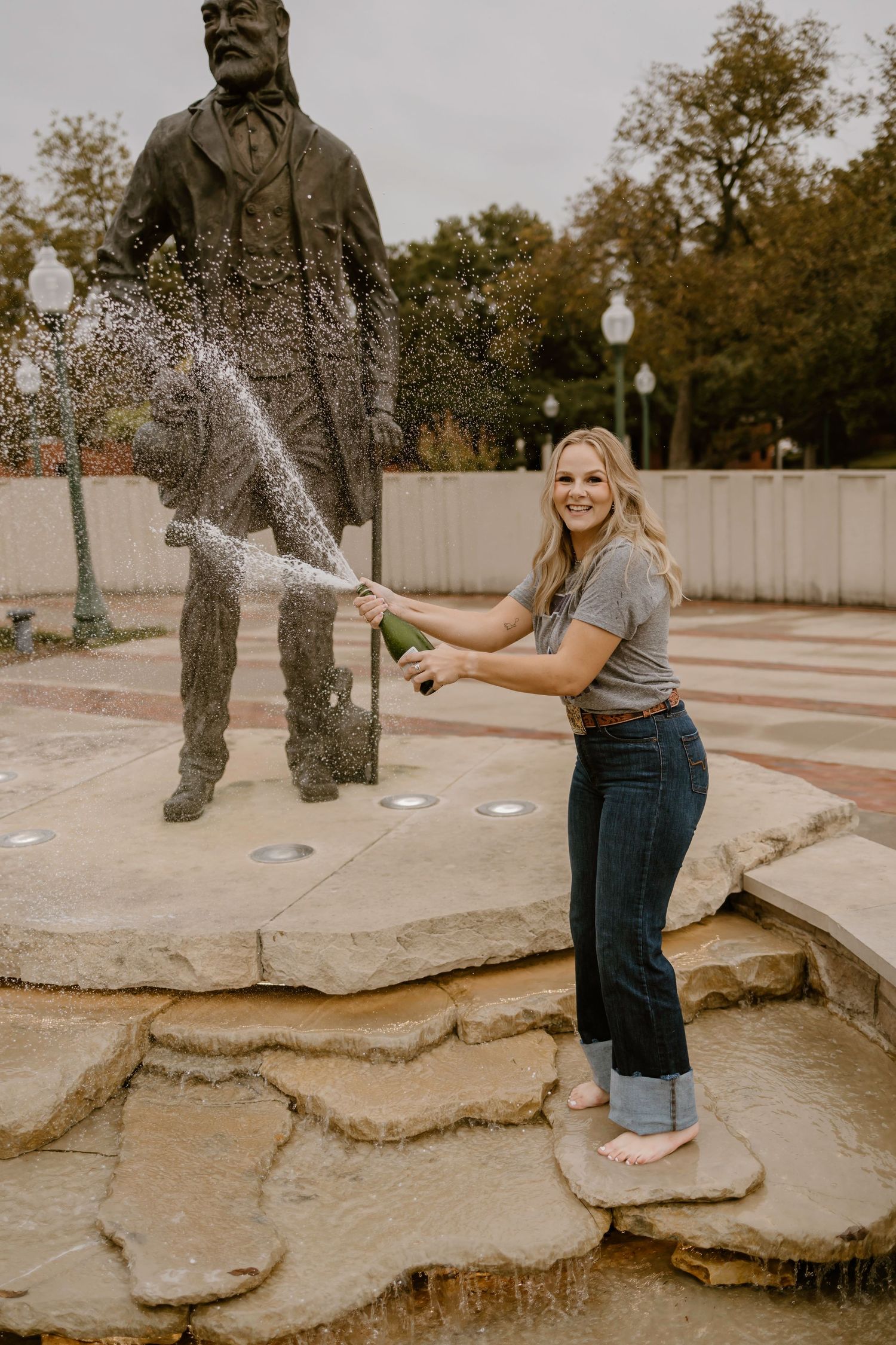 Water splashes dramatically from a bottle against a bronze statue in an outdoor fountain setting.