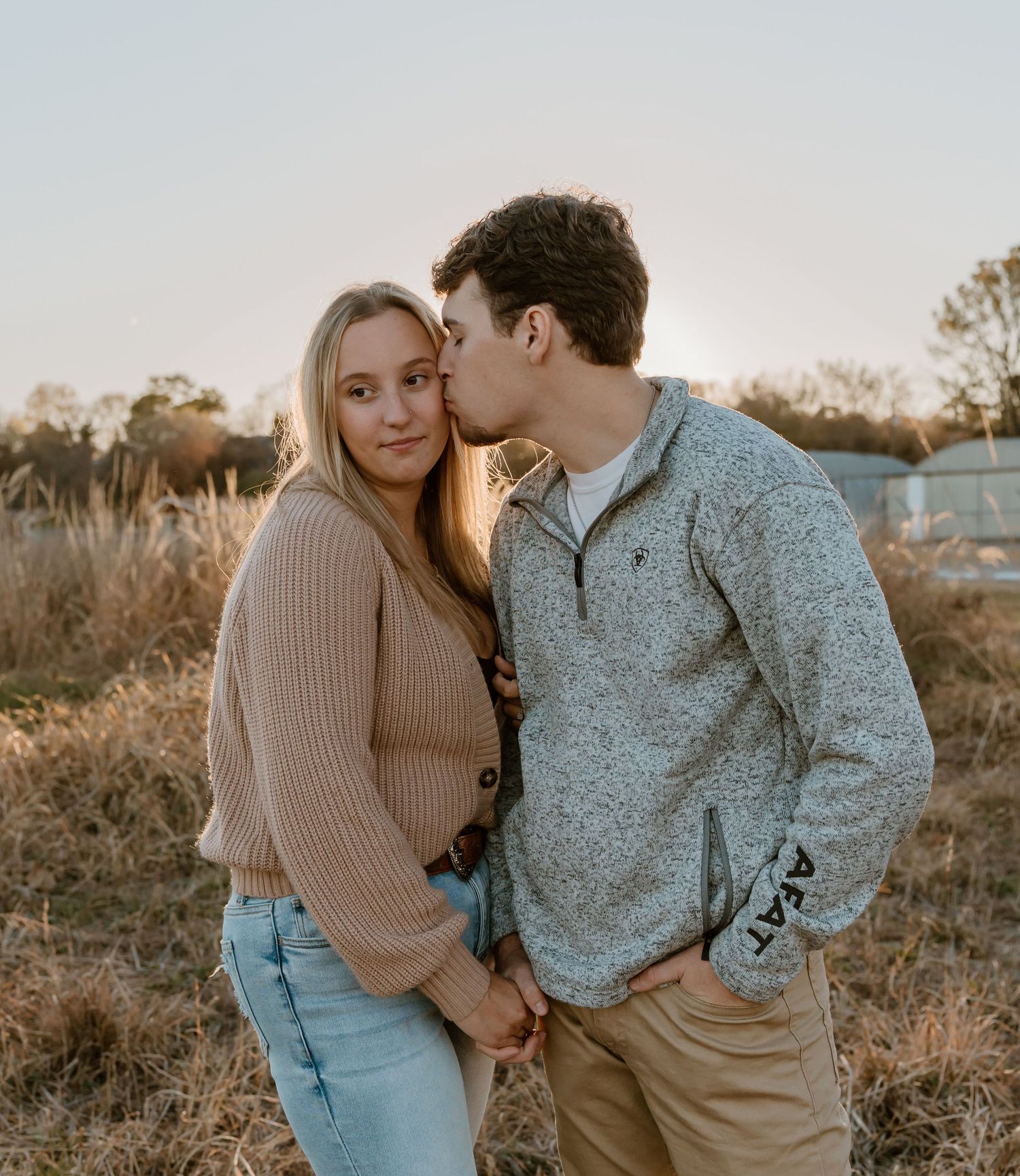 A romantic couple shares a tender moment at sunset in a field with tall grass and warm lighting.