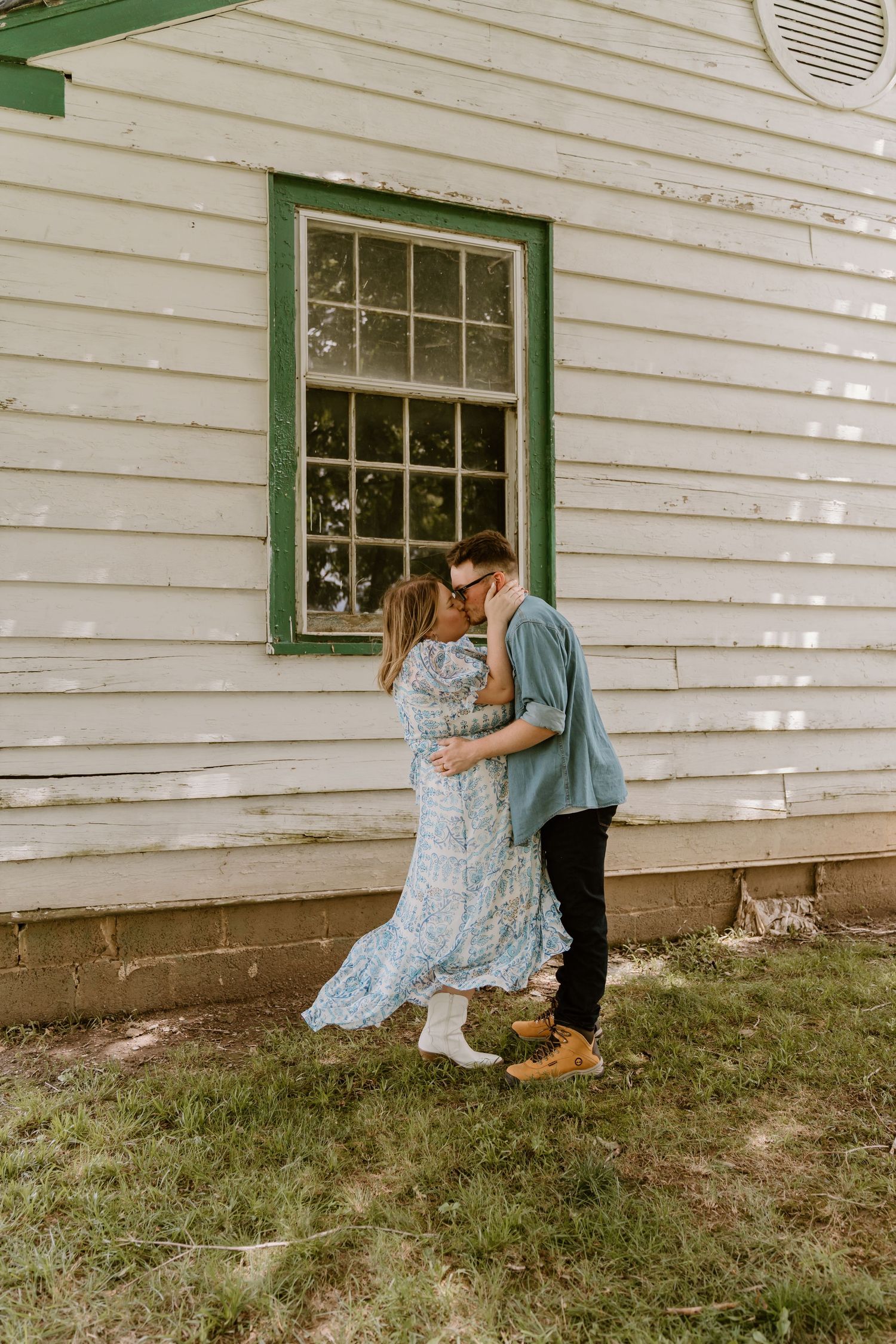 A couple shares a tender moment against a white farmhouse wall with green trim.