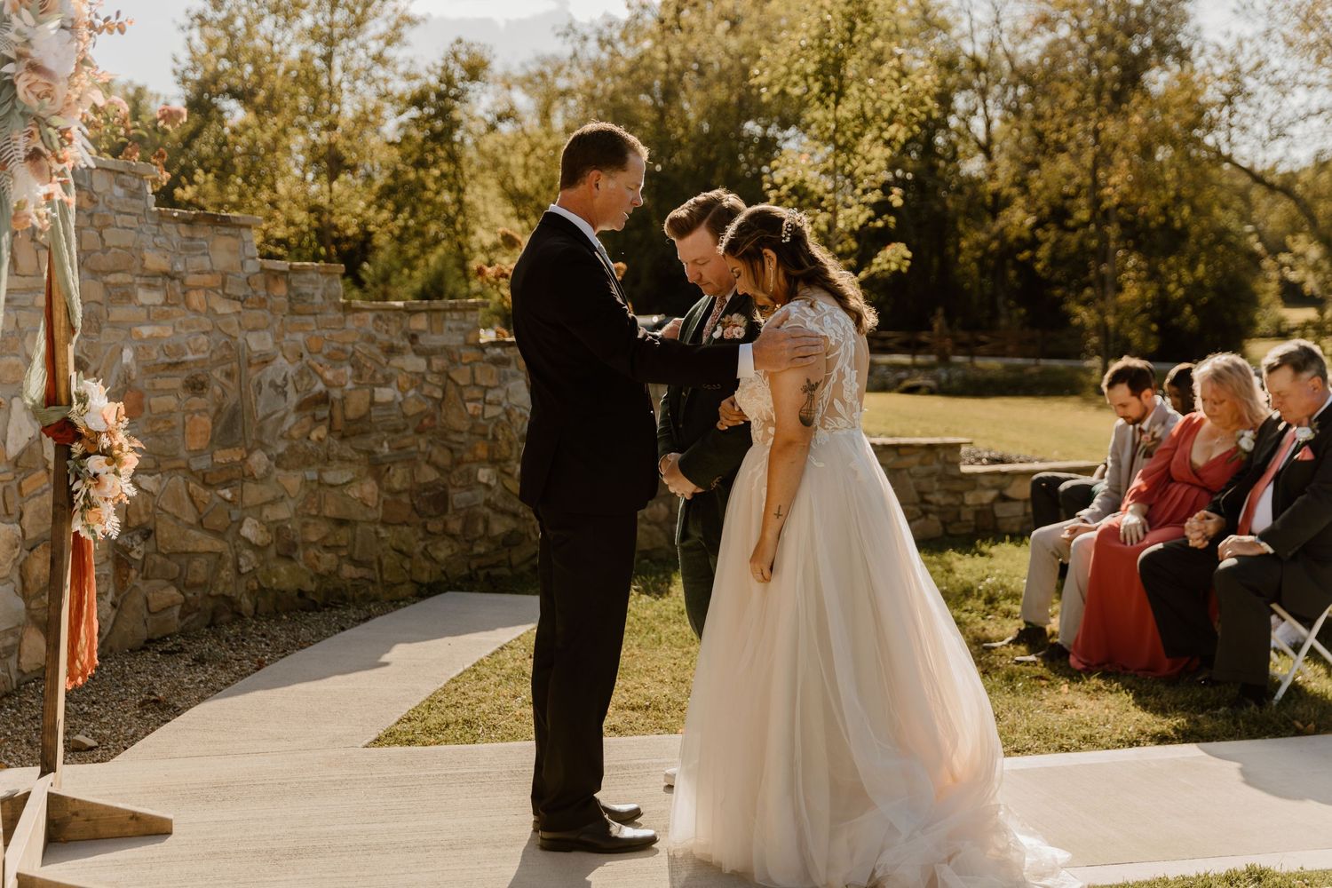 An outdoor wedding ceremony takes place against a rustic stone wall backdrop.