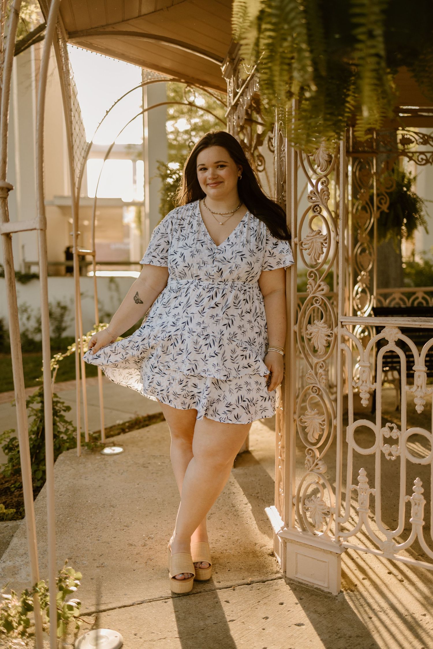 Person in floral dress poses elegantly on porch with ornate iron railings at sunset.