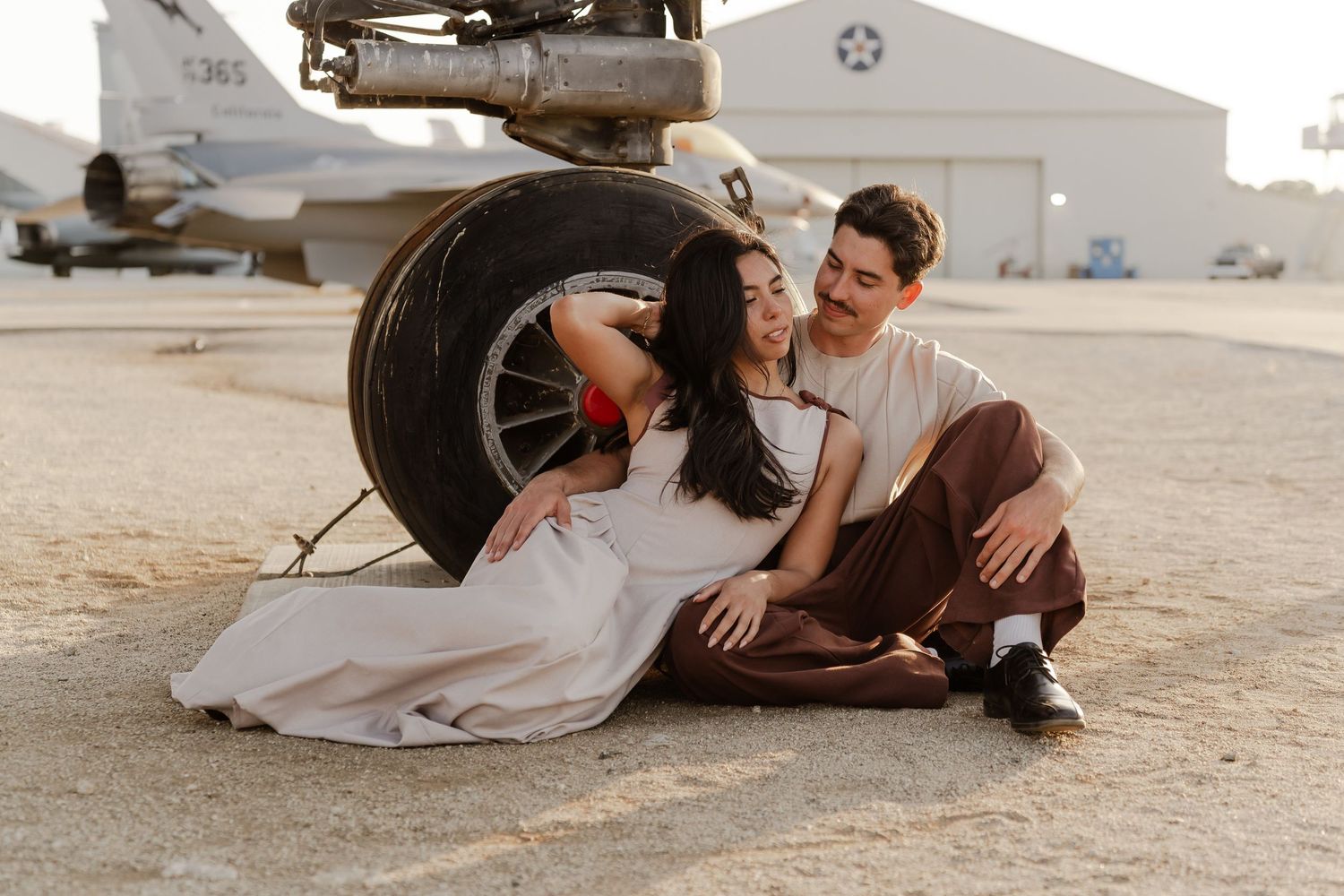 Couple sitting together on tarmac leaning against airplane landing gear at an airfield in a romantic engagement photo.