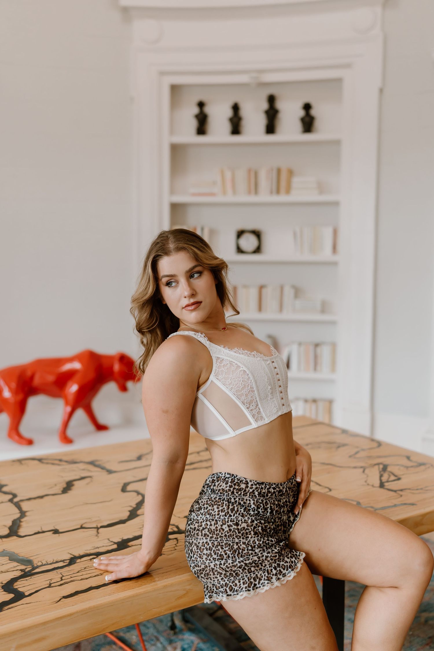 Woman in white lace bralette and leopard print shorts posing by a wooden table in a bright room.