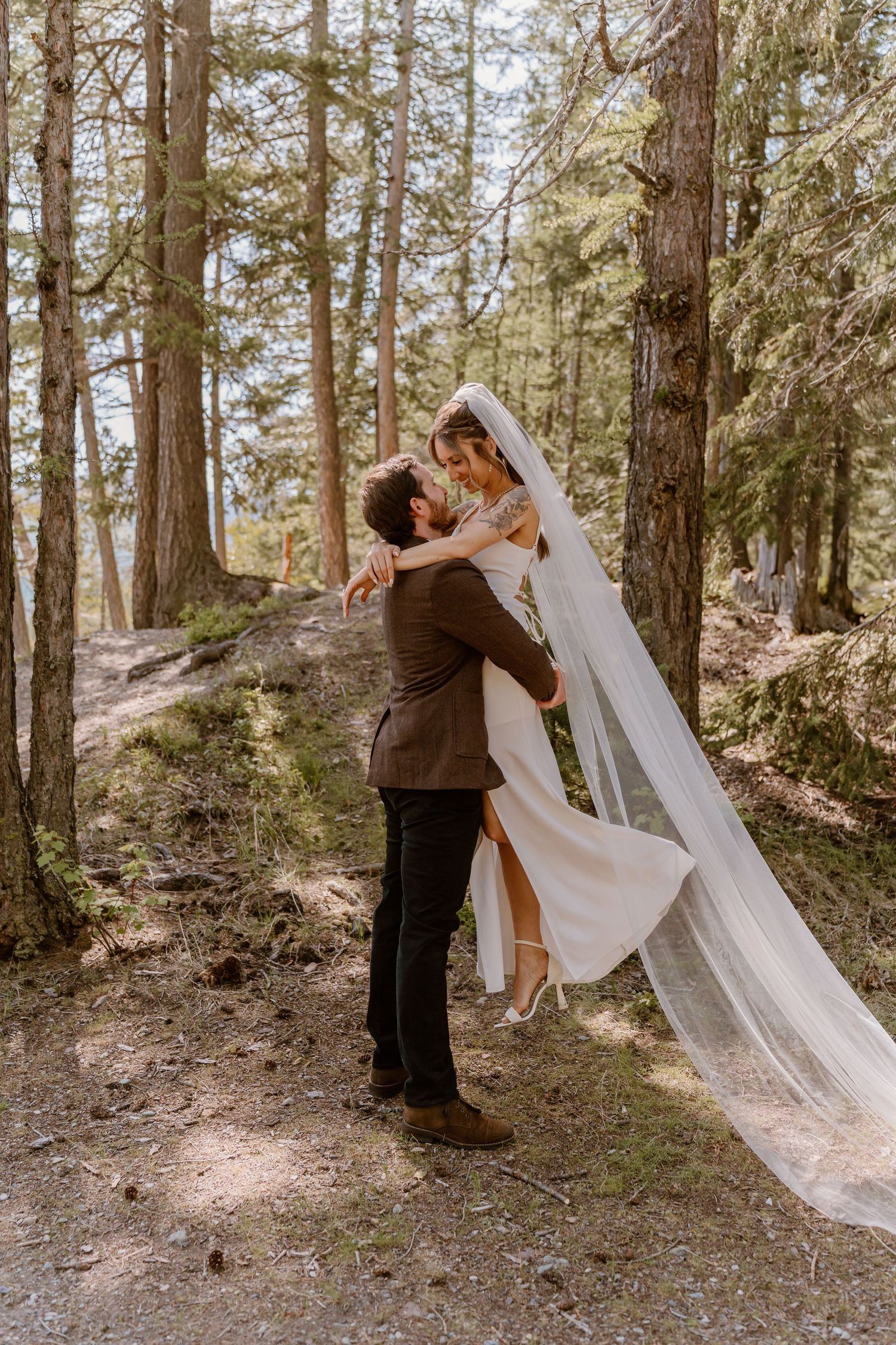 A romantic wedding couple embraces in a sun-dappled forest setting while the bride's veil flows gracefully in the breeze.