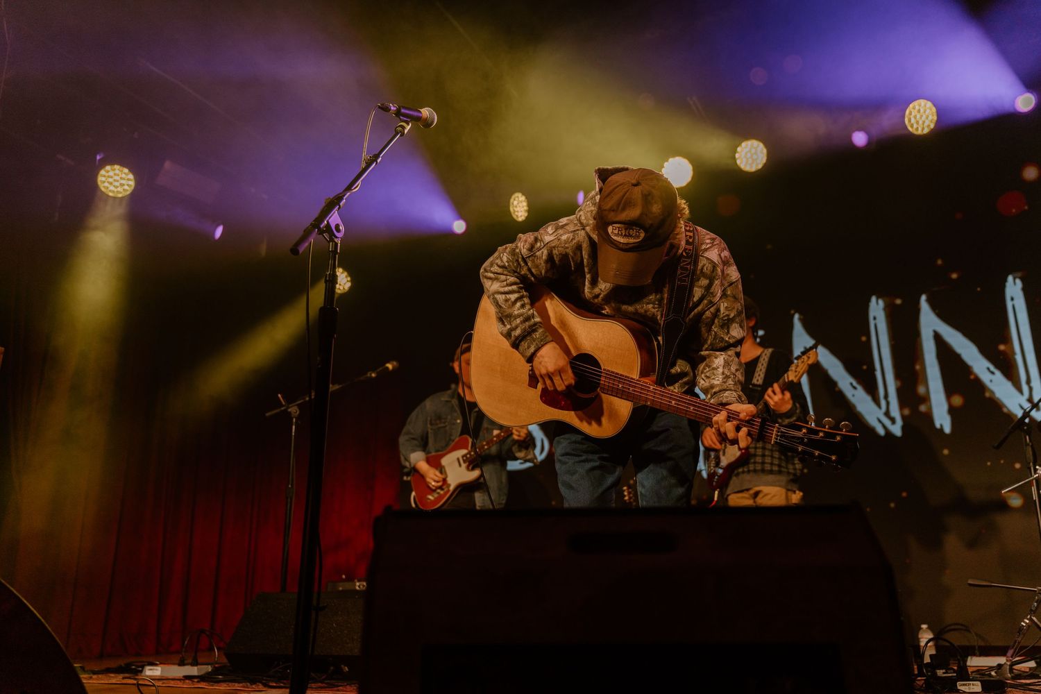 Musicians performing on stage with purple ambient lighting and concert equipment visible.