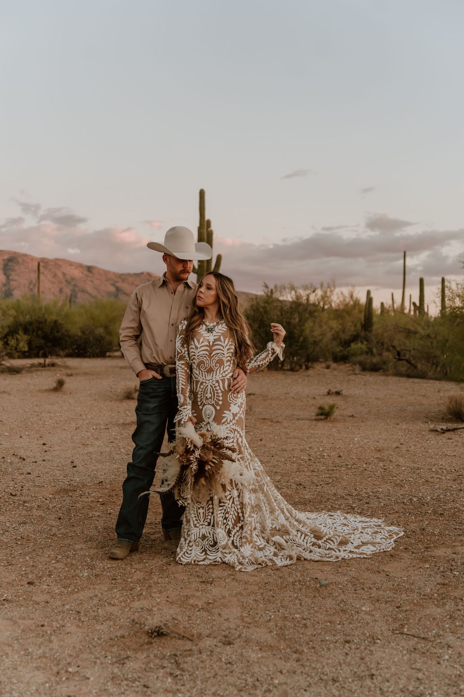 Wedding couple poses in desert landscape with dramatic mountain backdrop and saguaro cacti.
