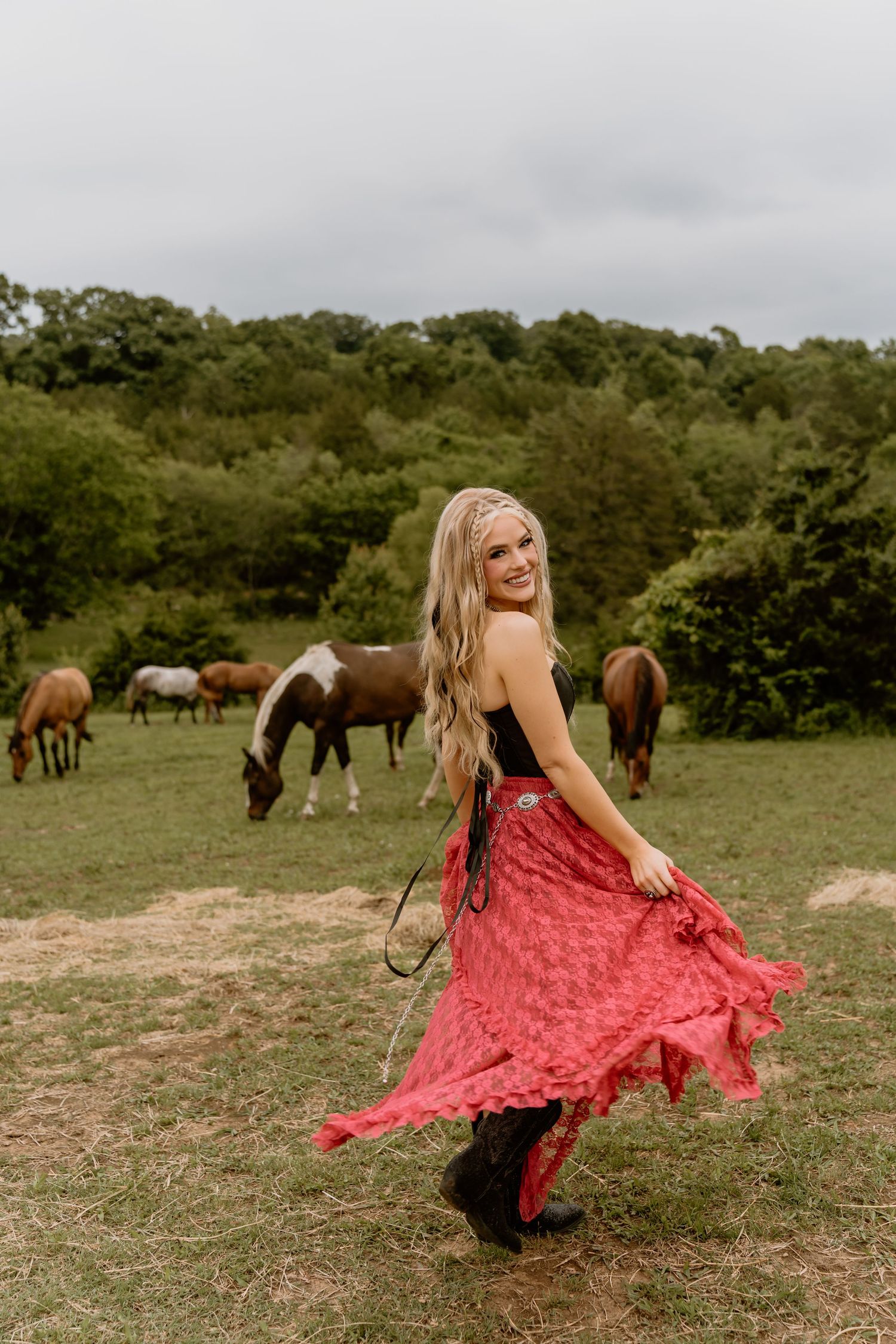 Woman in flowing red dress dances playfully in a field with horses grazing in the background.