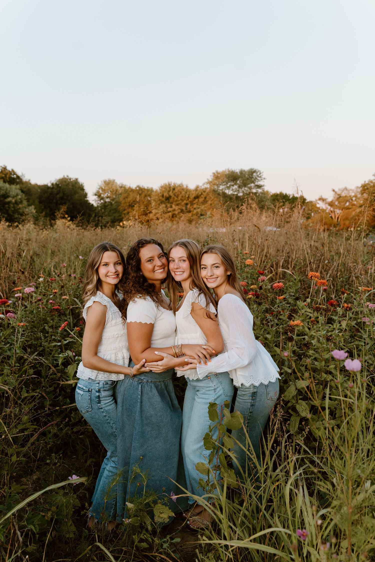 Friends in white tops and jeans pose together in wildflower field at sunset.