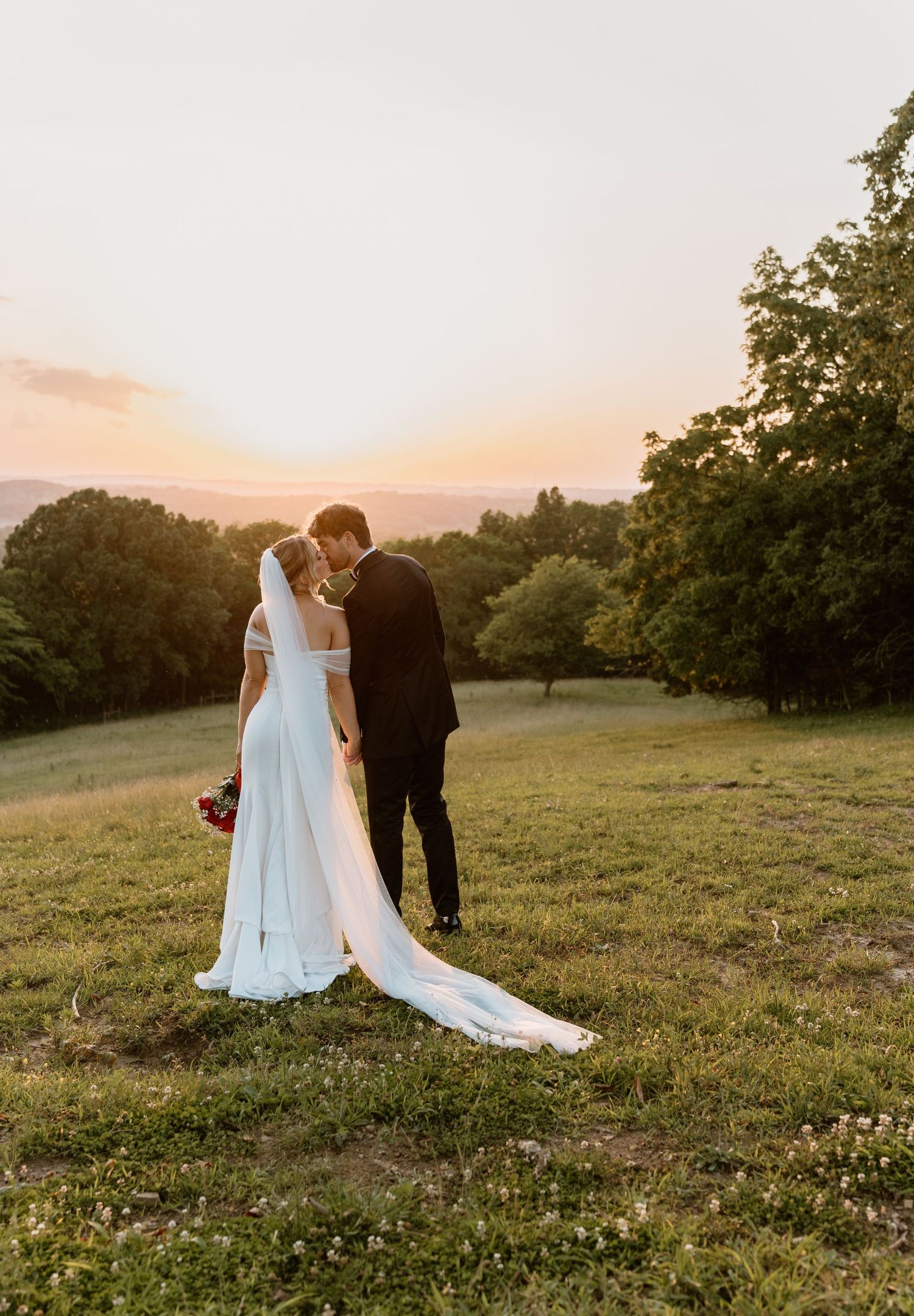 A couple shares a romantic moment at sunset on a hilltop meadow, with the bride's white dress flowing in the golden light.