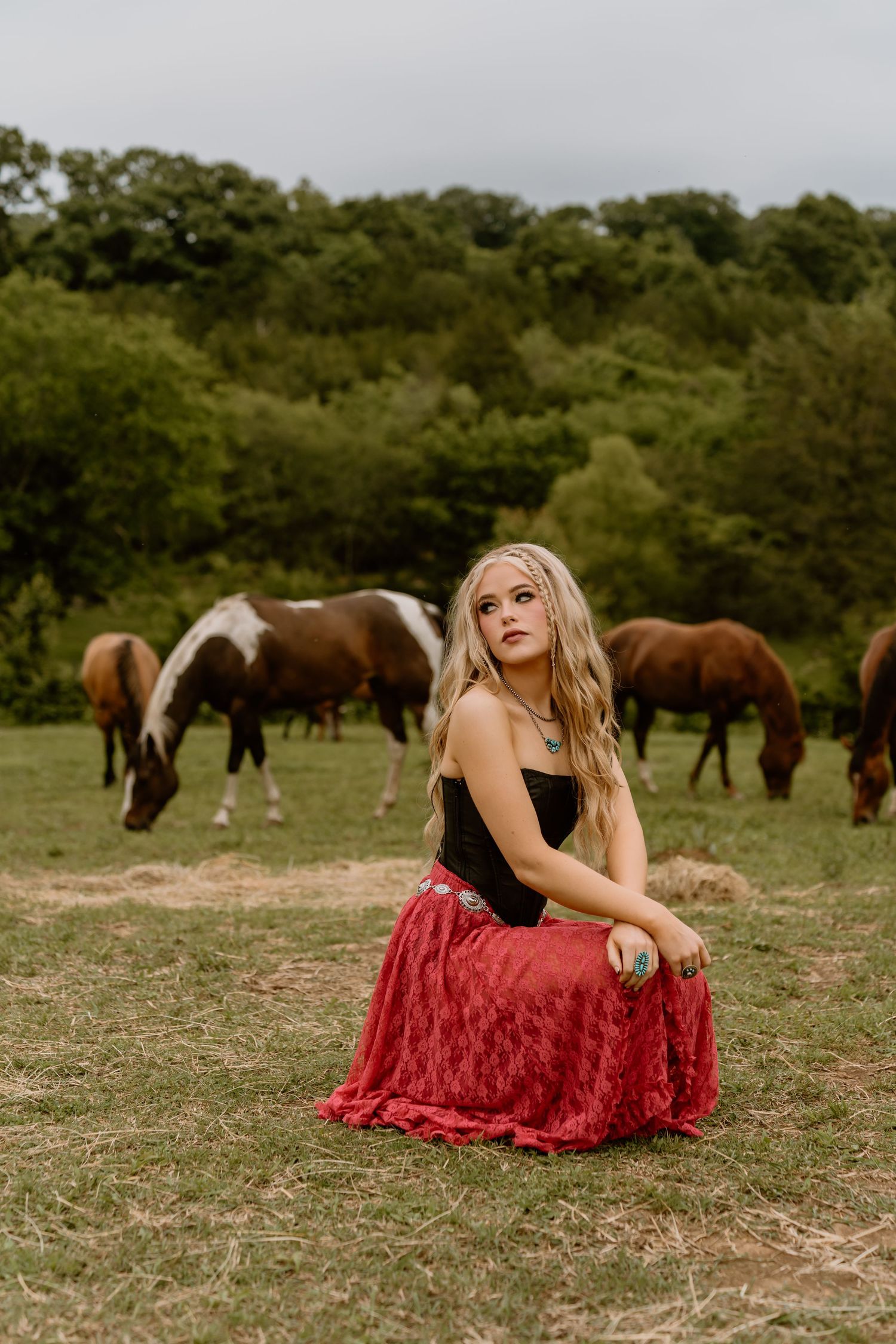 Red dress contrasts against a pastoral field with horses grazing in the background.