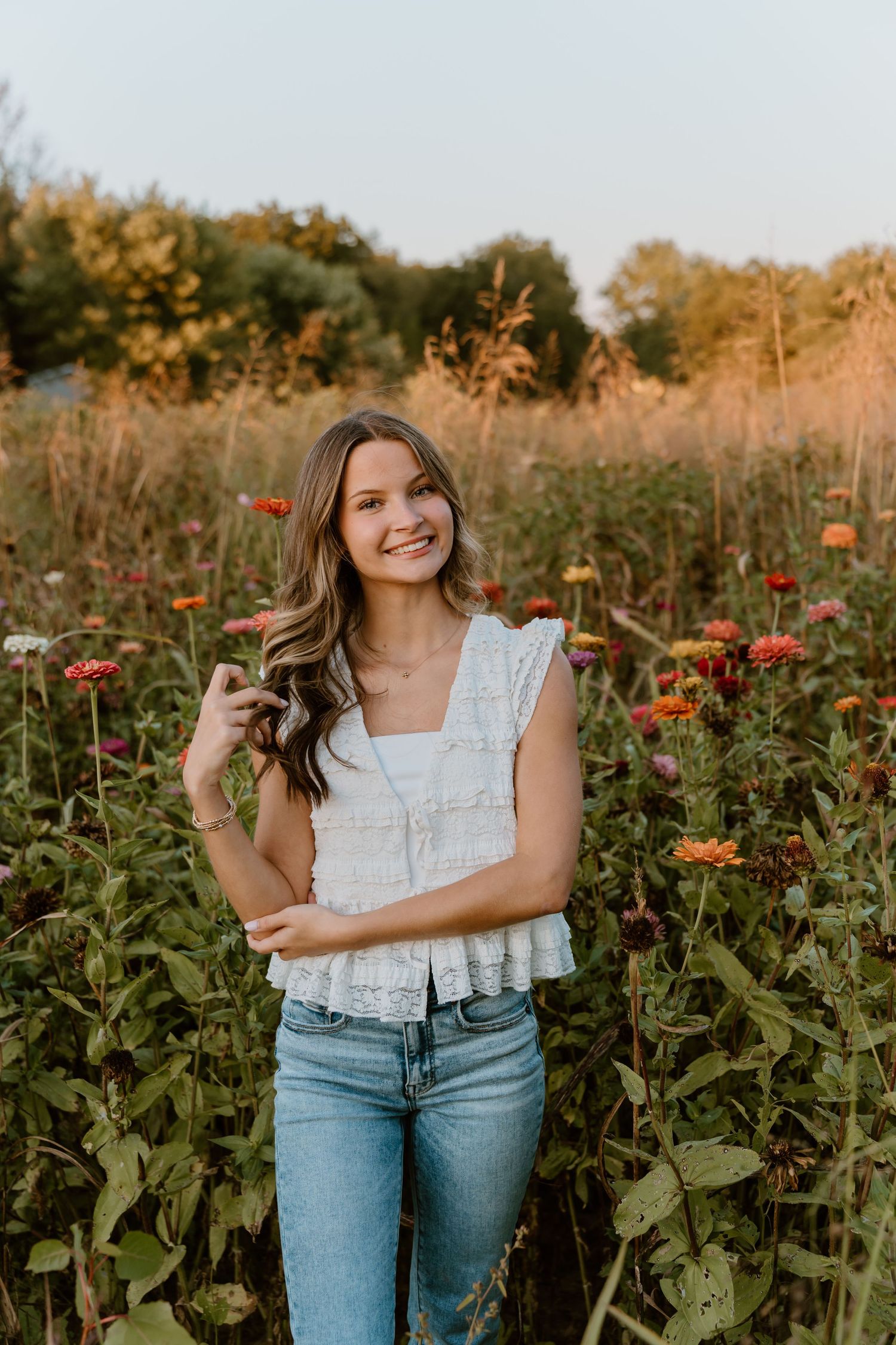 Woman in white ruffle top and jeans poses in garden of blooming red flowers.