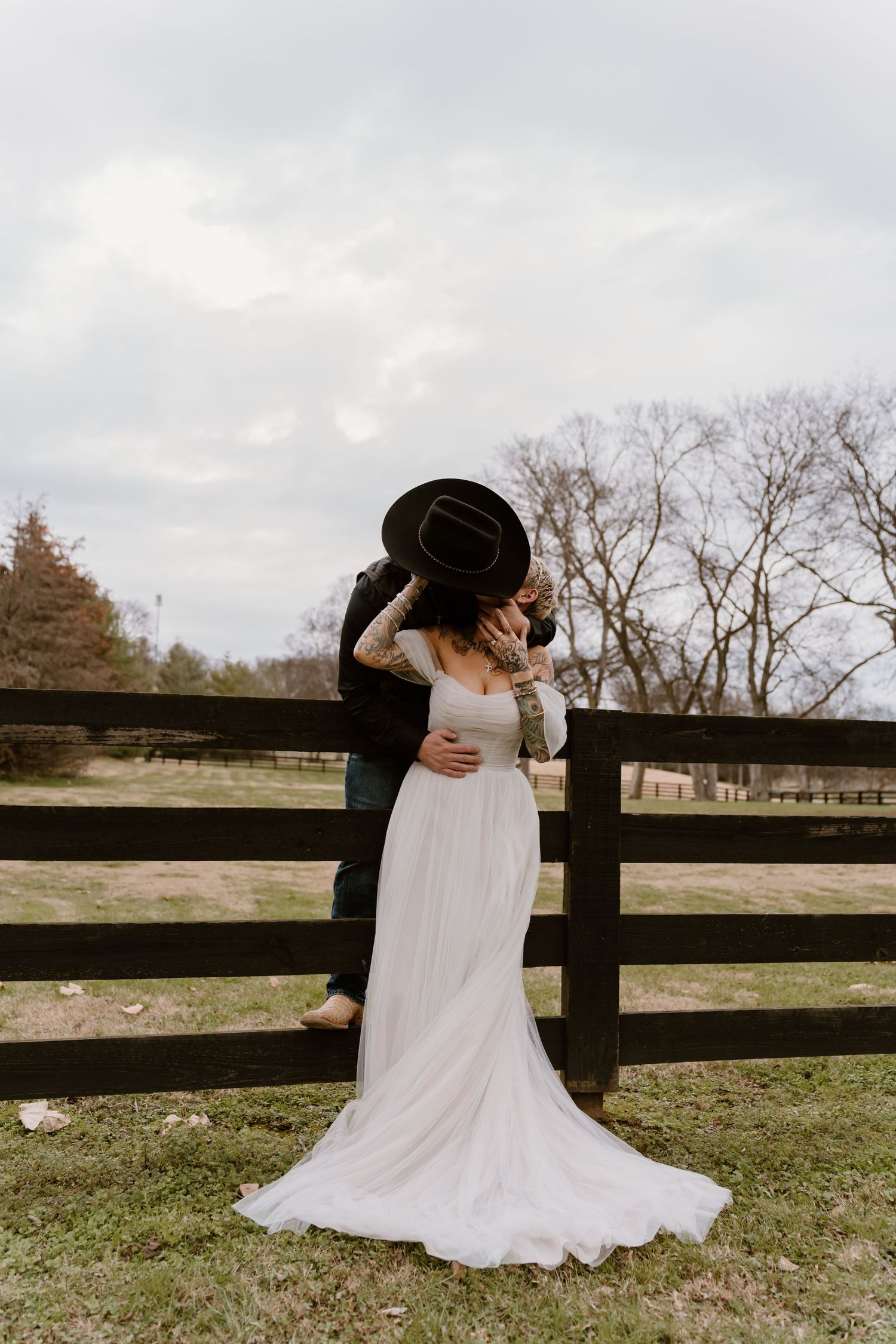A romantic outdoor wedding scene with a couple embracing by a rustic wooden fence in a pastoral countryside setting.