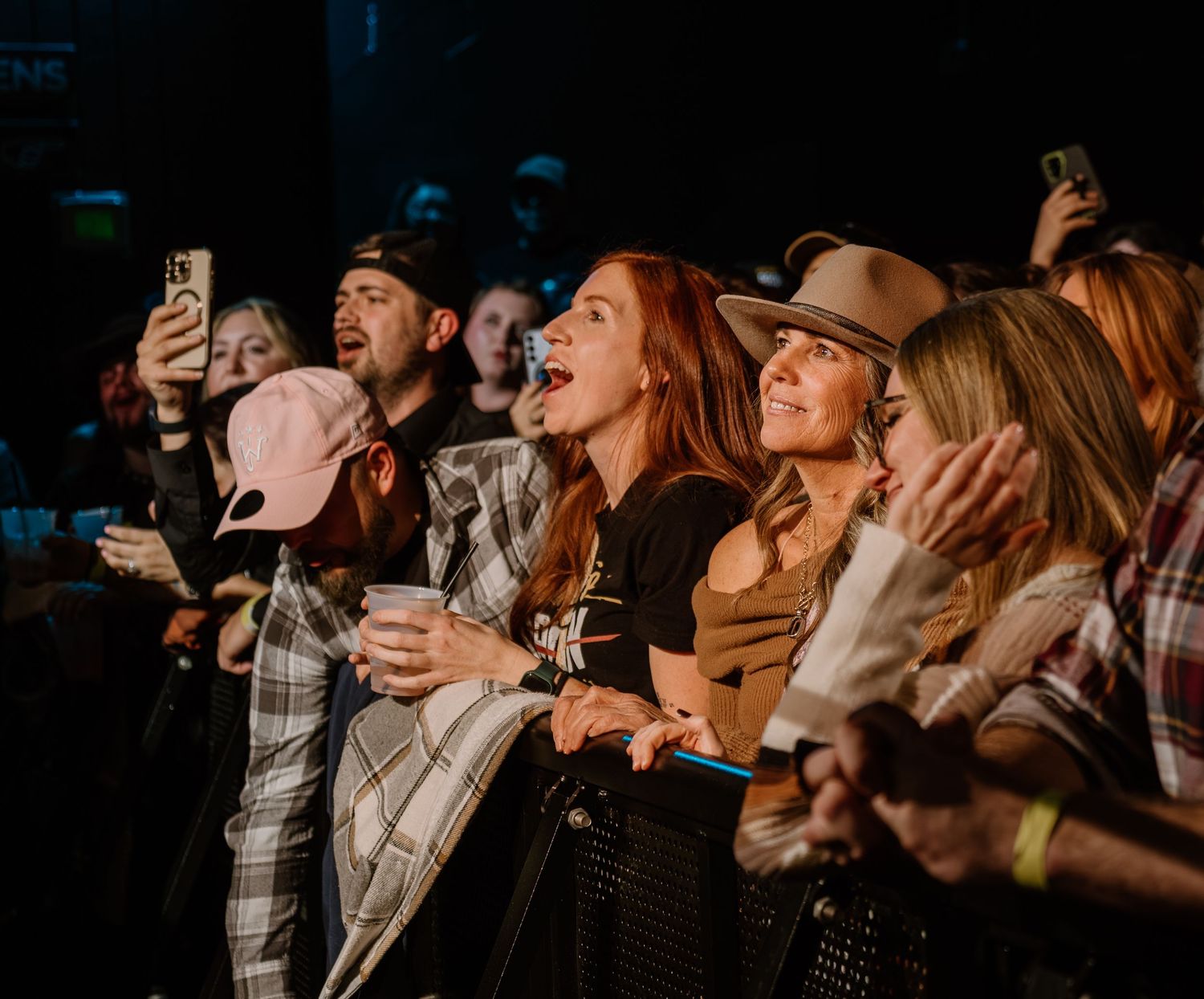 Concert crowd cheering and singing along enthusiastically at a dimly lit music venue.