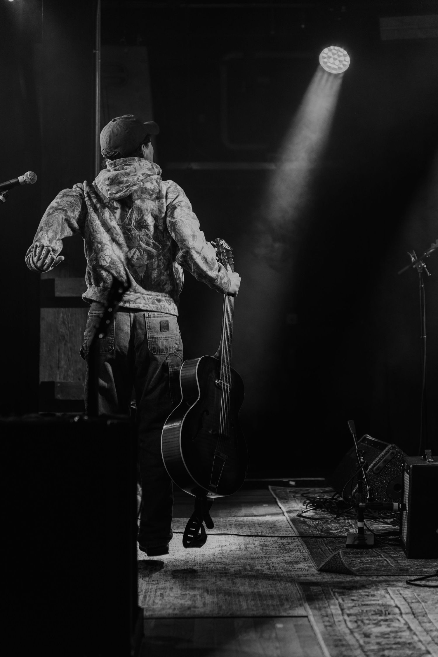 Black and white photo of a guitarist holding an acoustic guitar on a dimly lit concert stage with spotlight.