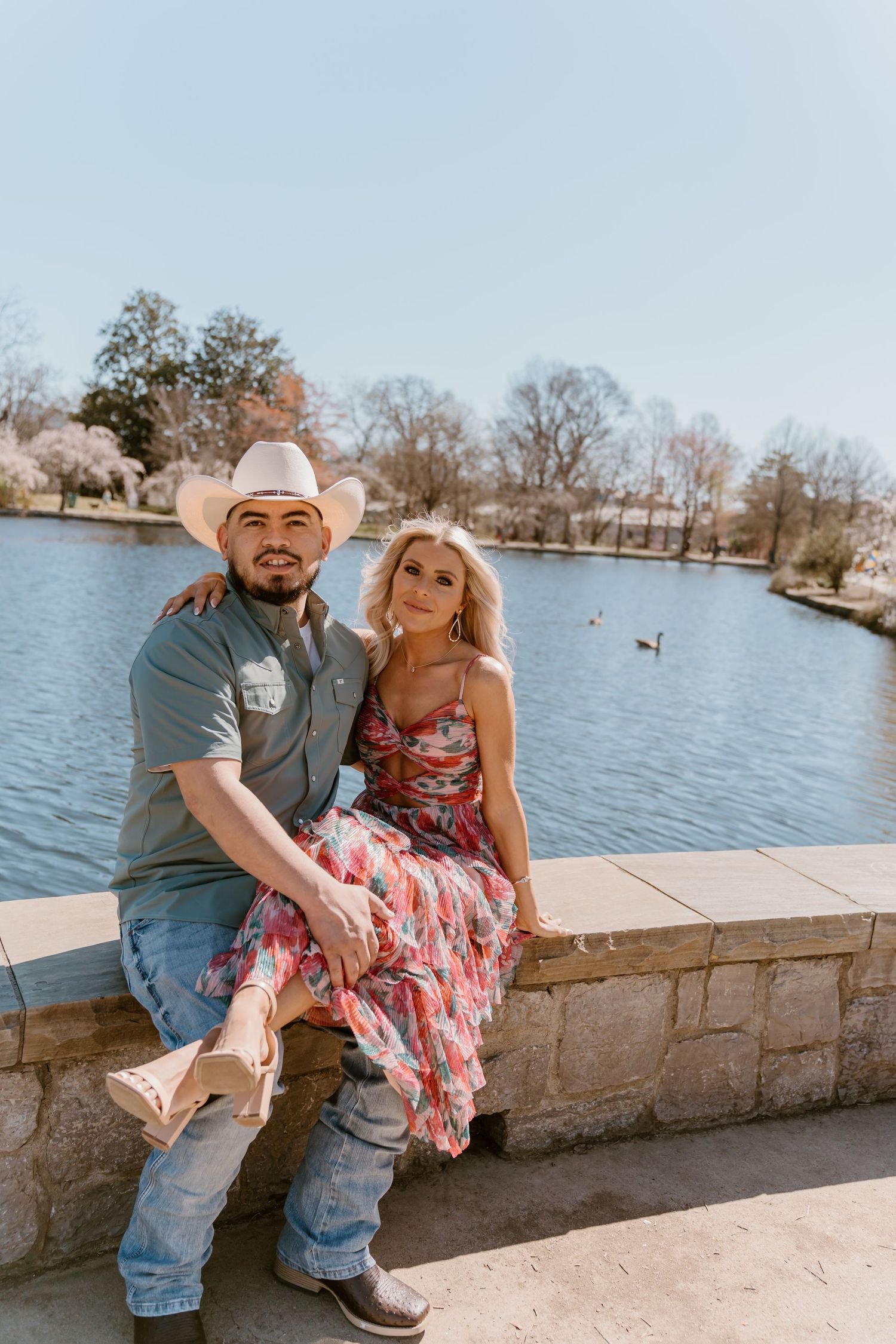 A couple sits together on a stone wall by a lake with ducks swimming in the background.