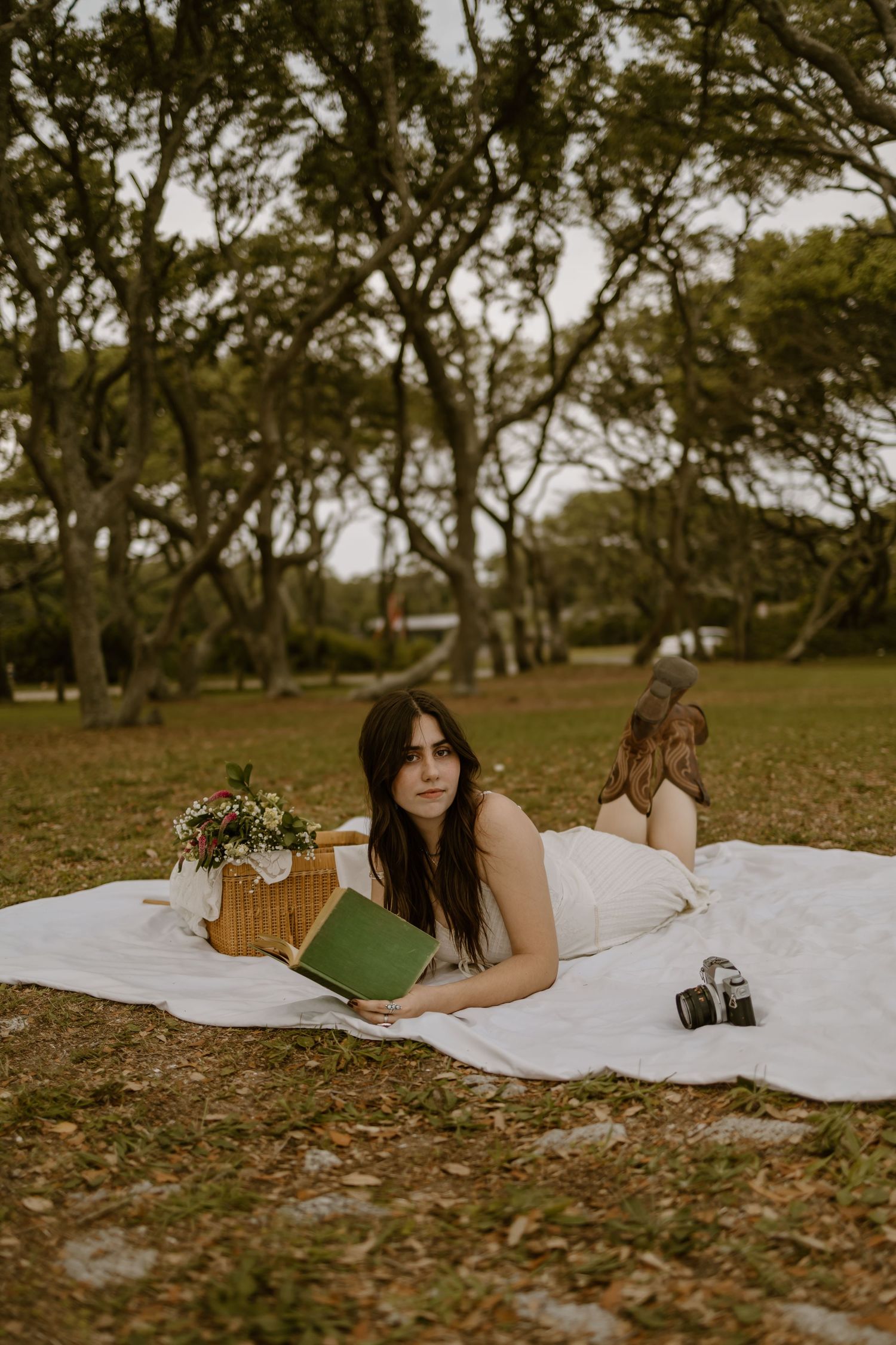 Two people enjoying a picnic on white blanket with books and flowers under shady trees.