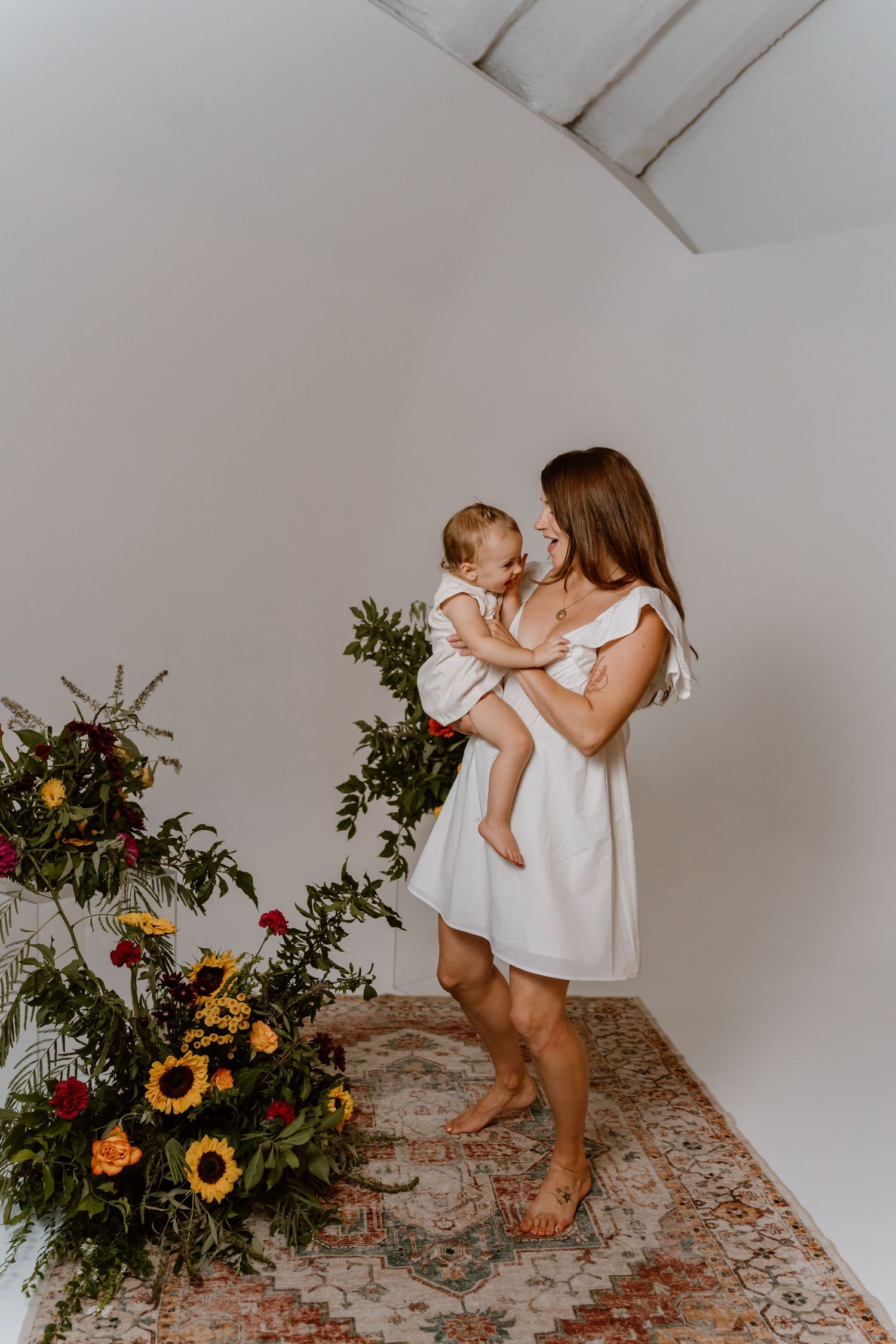 Parent and child share tender moment in white room with floral arrangements.