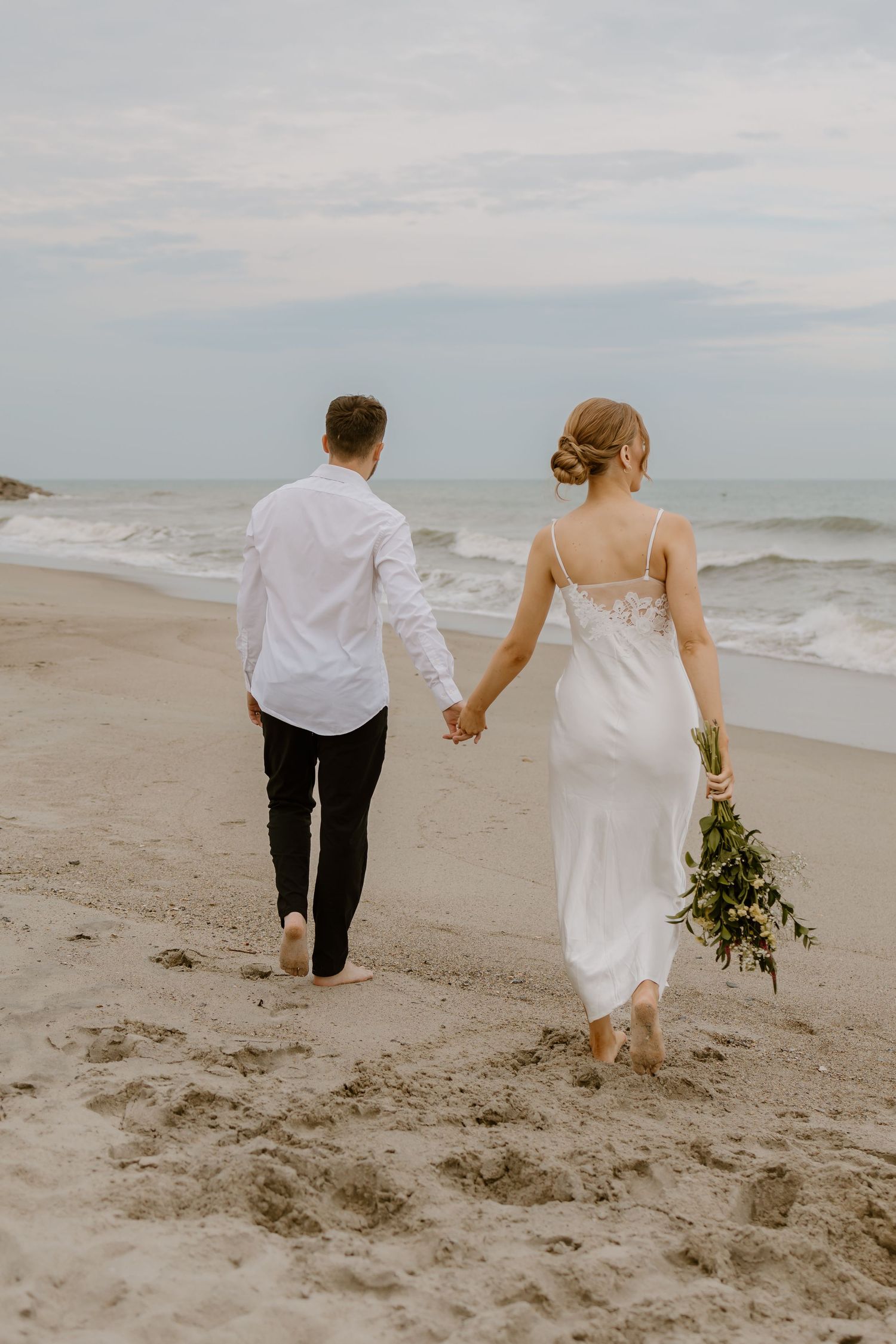 A couple walking hand in hand along a misty beach shore in wedding attire.