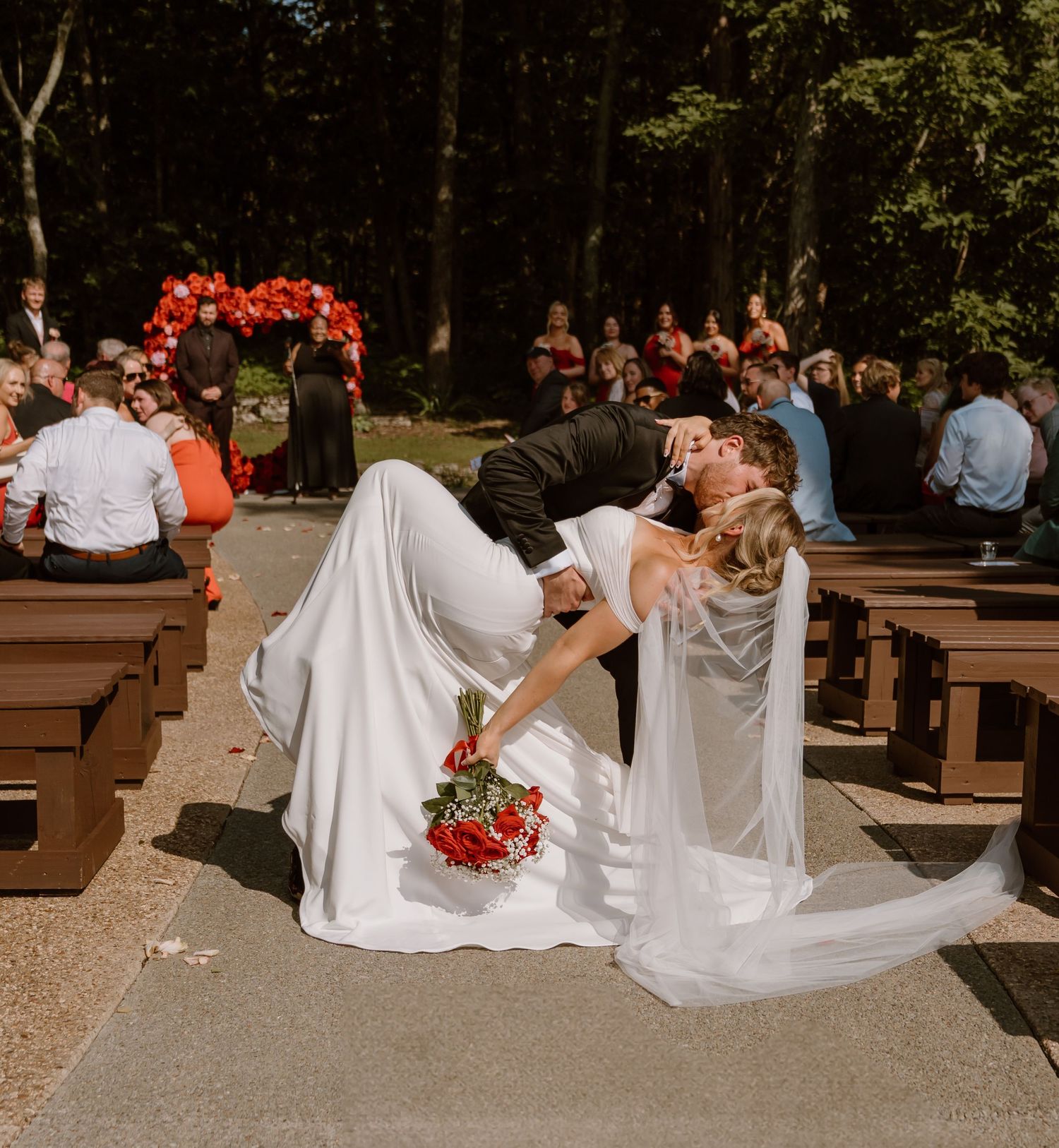 A wedding couple sits on wooden benches in an outdoor forest setting surrounded by guests during a ceremony.