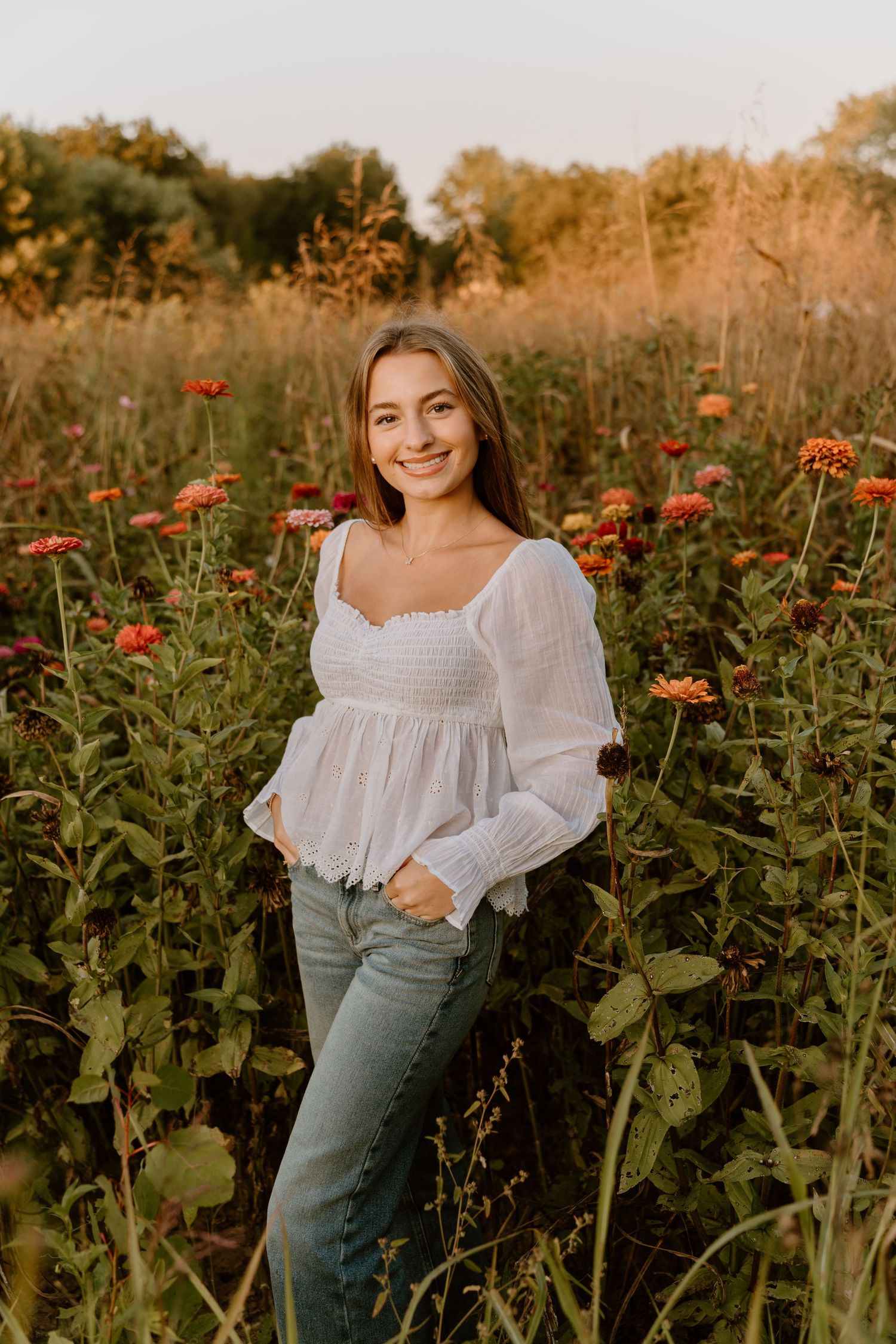 A woman in a white peasant top poses in a field of orange zinnias during golden hour.