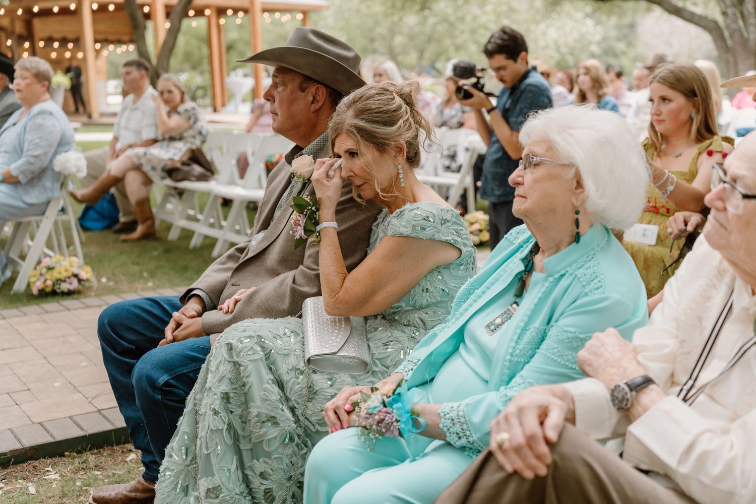 Emotional wedding guests seated outdoors, woman wiping tears during ceremony at rustic garden venue.