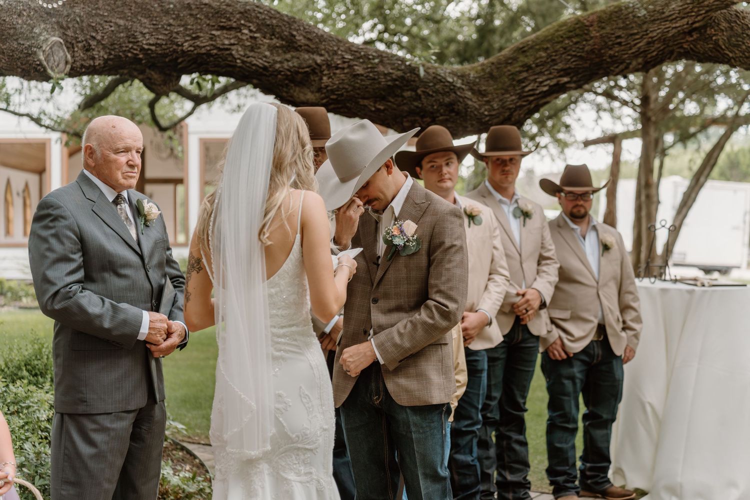 Rustic outdoor wedding ceremony with groom in cowboy hat, groomsmen in jeans, and bride in white gown under oak tree.