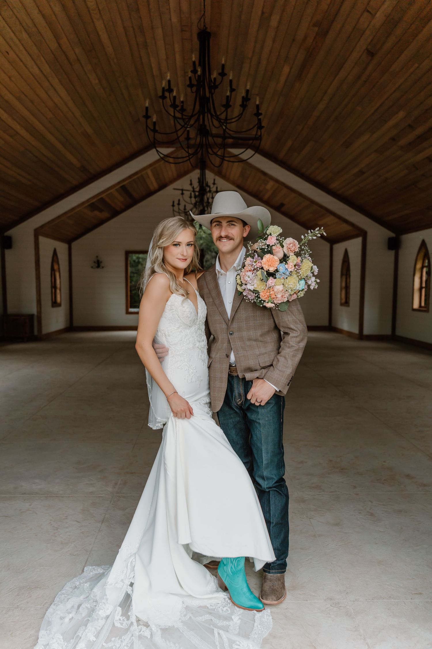 Bride in white lace gown and groom in cowboy hat with turquoise boots pose in rustic chapel holding floral bouquet.