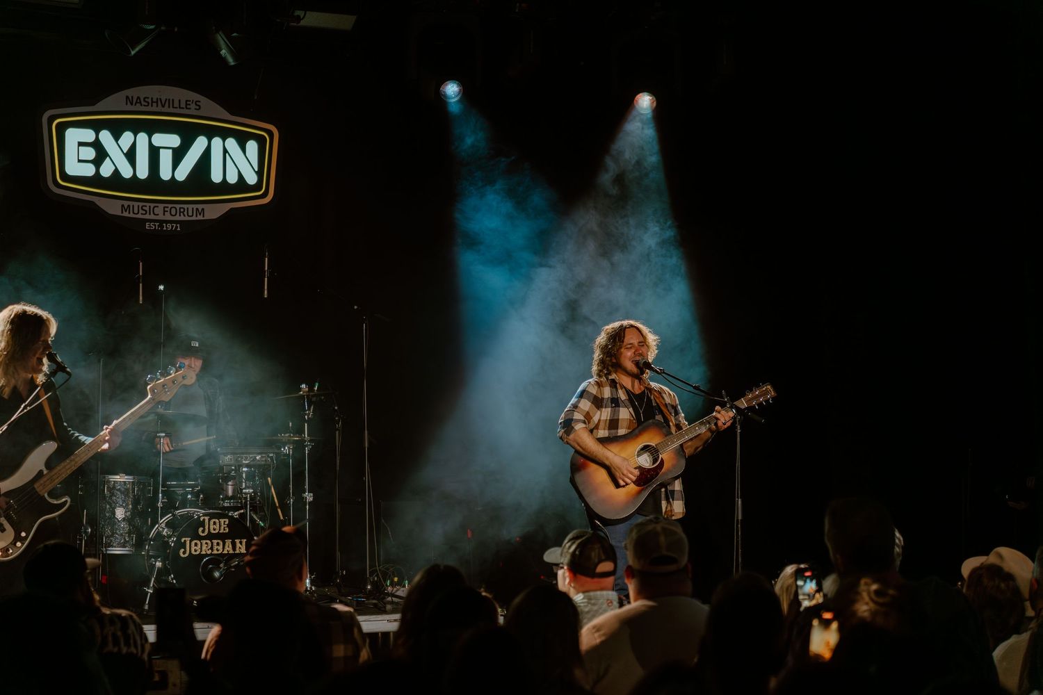 Musicians perform on a dimly lit stage with dramatic blue lighting and fog effects during a live concert performance.
