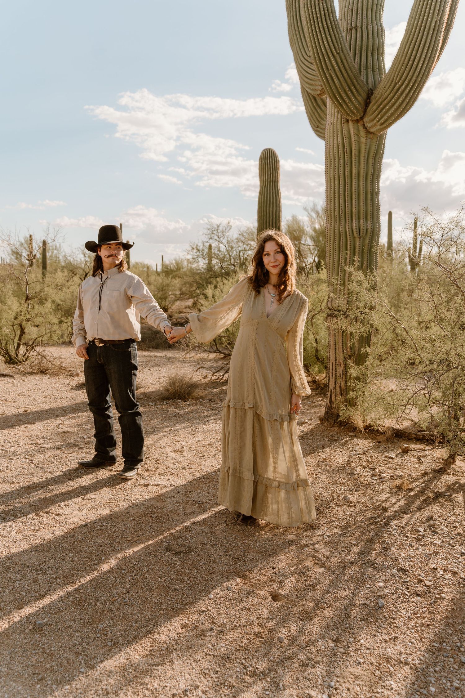 A couple stands together in a desert landscape with towering saguaro cacti against a bright sky in Arizona.