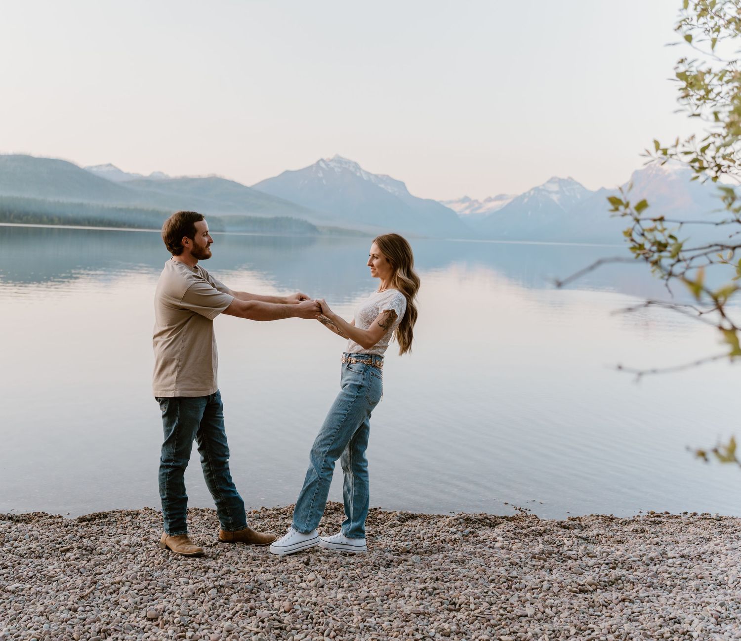 A couple stands at the edge of a rocky beach with mountains and a serene lake in the background during sunset.