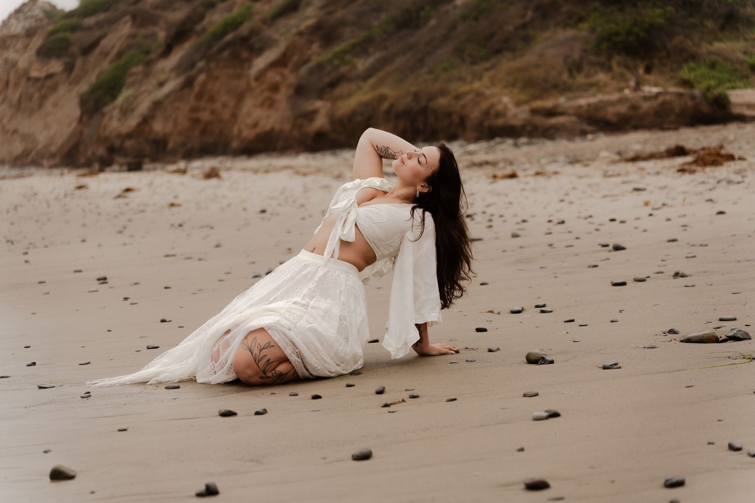Woman in white dress poses gracefully on sandy beach, leaning back with hand in hair, rocks and cliffs in background.