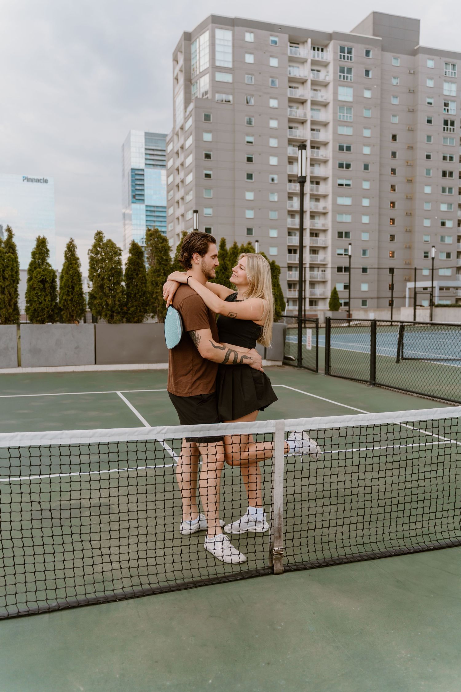 A playful couple sits on a tennis court net with city buildings in the background.