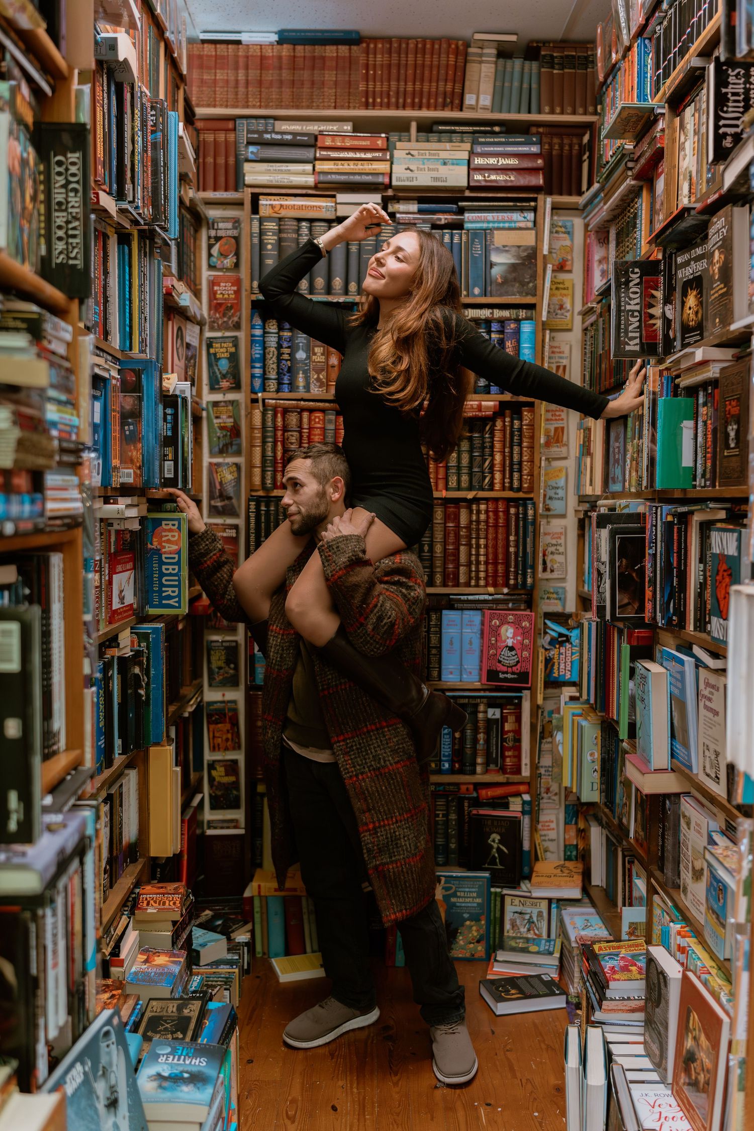Two people pose playfully between tall bookshelves in a cozy bookstore filled with vintage and classic books.