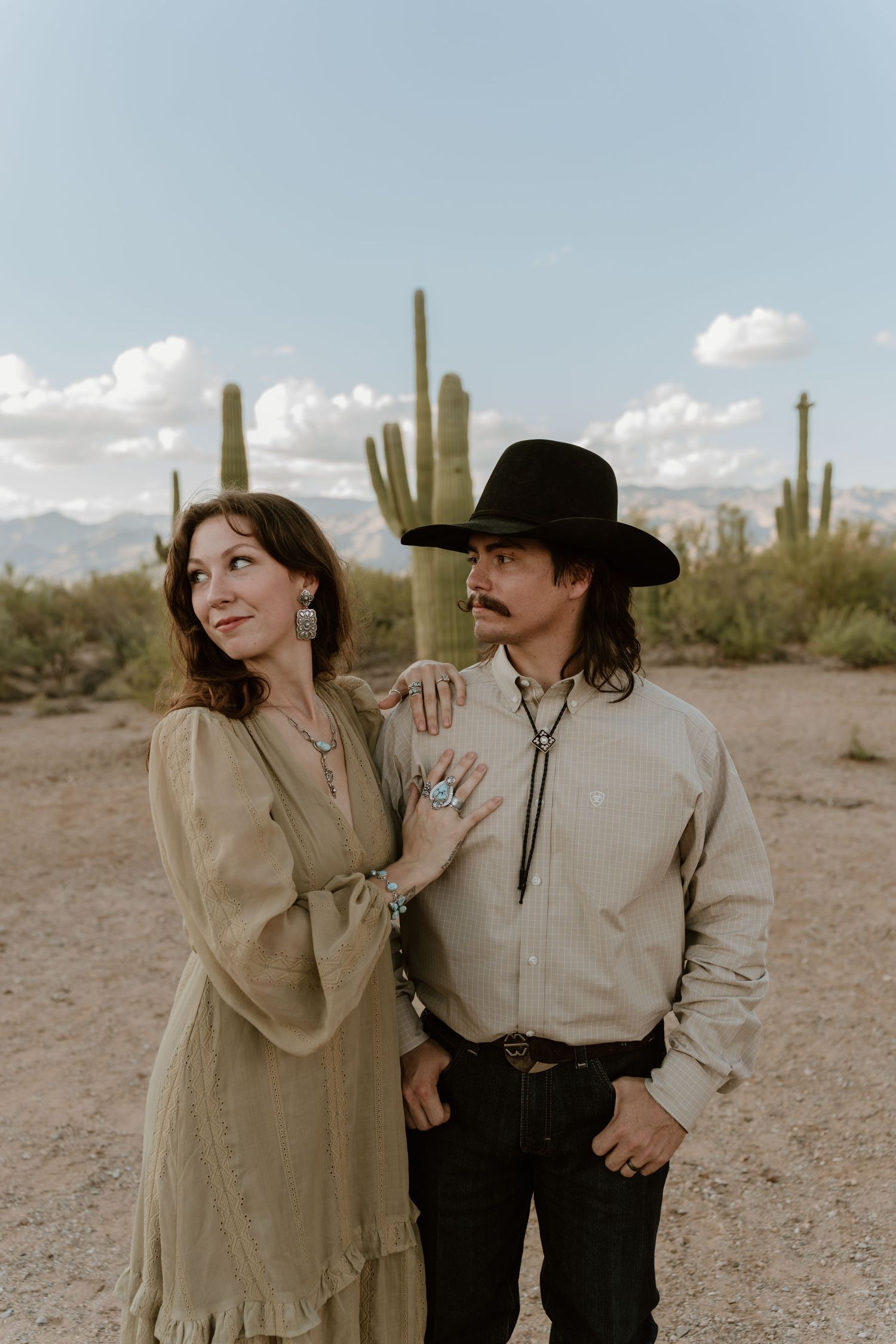 A couple poses together in a desert landscape with saguaro cacti and mountains in the background.